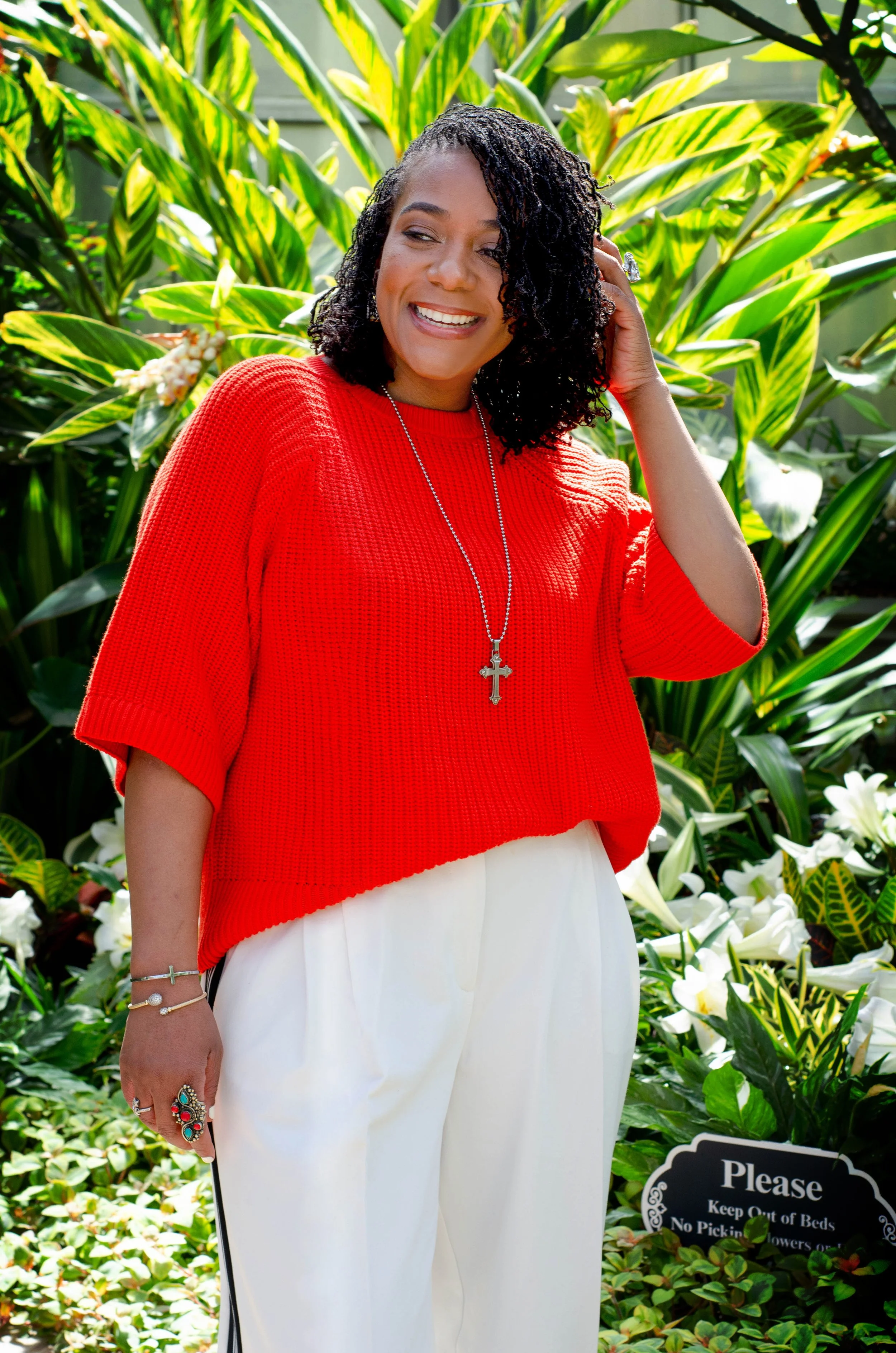 A woman smiling in a garden with large green leafy plants behind her. She is wearing a red top, white pants, and jewelry, including a cross necklace and rings.