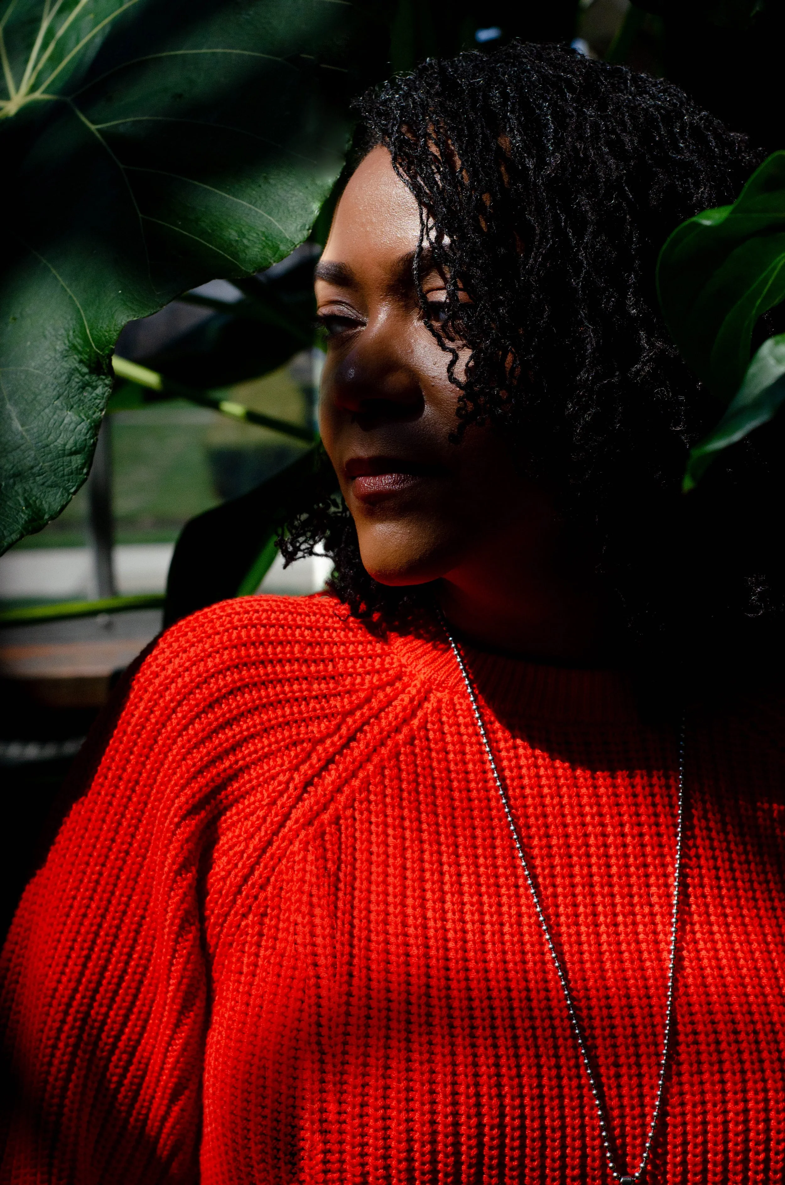 A woman with brown skin and dark black hair, wearing a bright orange knit sweater, is surrounded by large green leaves and appears to be in a lush, indoor greenhouse environment.