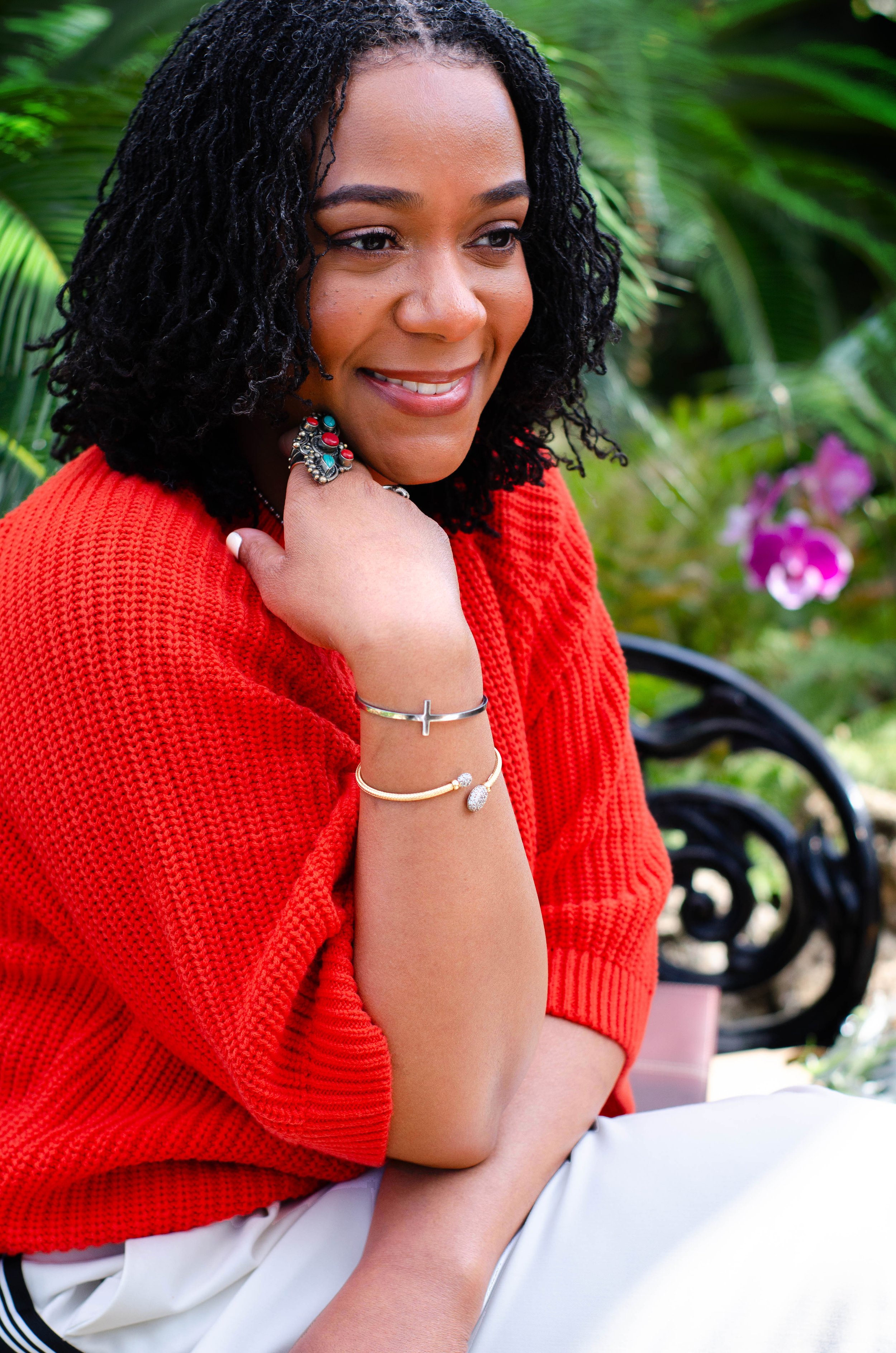 A woman with curly black hair and light brown skin wearing a red sweater, sitting outdoors in a garden with greenery and purple flowers in the background, smiling and resting her chin on her hand.