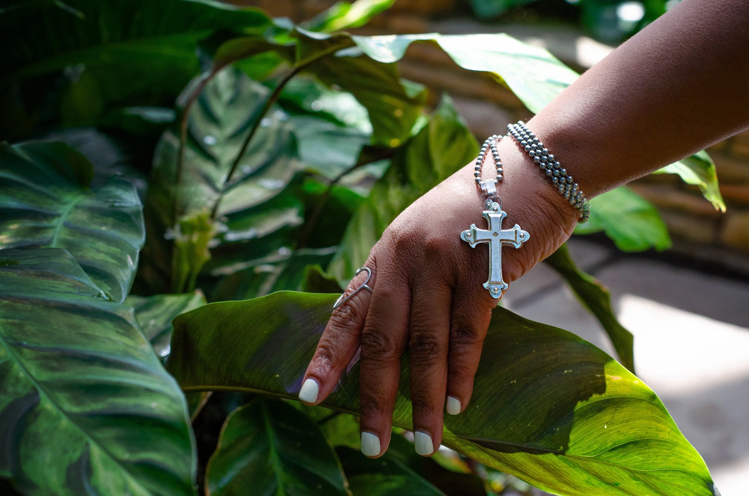 A person's hand with white-painted nails and a silver cross bracelet is resting on green houseplant leaves, with a silver rosary bracelet hanging from the wrist, in a sunlit environment.