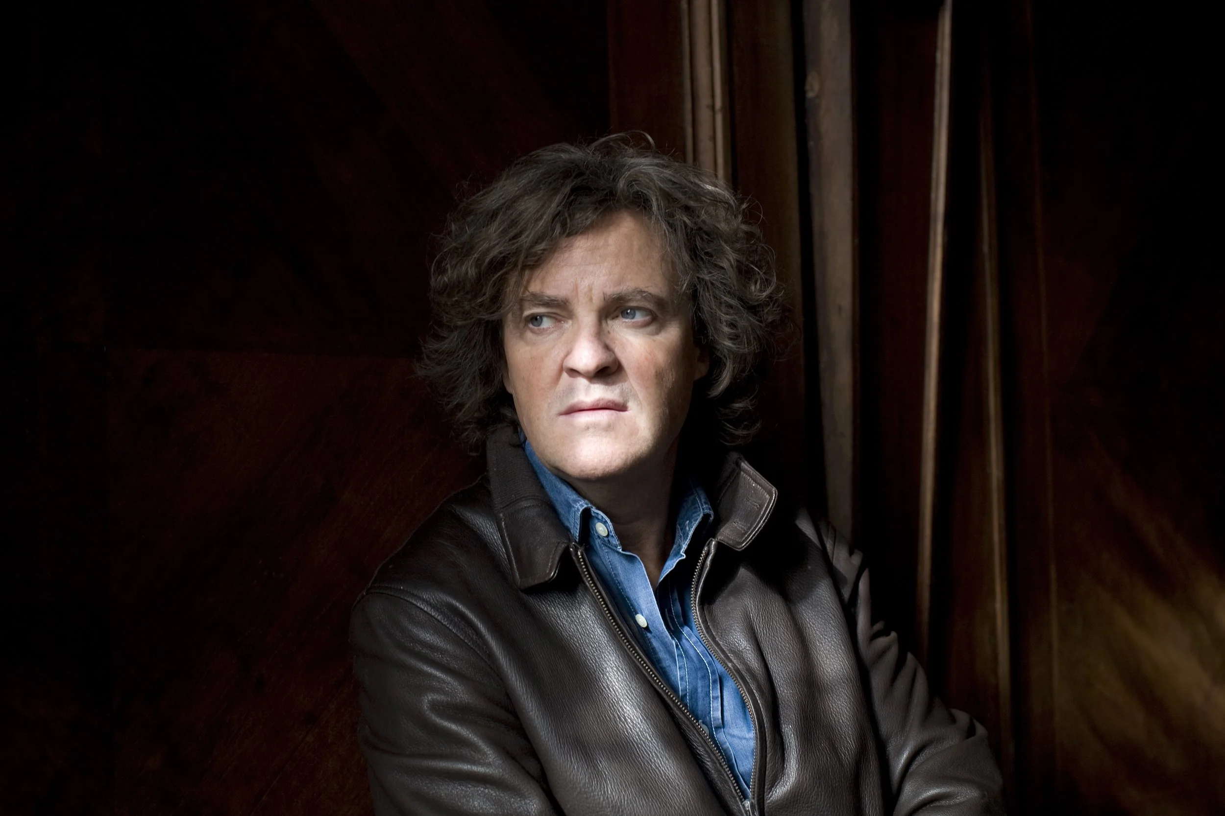 A man with curly brown hair and blue eyes wearing a black leather jacket and blue denim shirt, looking to the side against a dark wooden background.