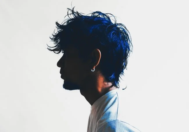 Silhouette of a man with spiky blue hair, wearing an earring, against a plain background.