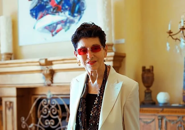 An elderly woman with short dark hair, wearing red-tinted sunglasses, a white blazer, a black patterned blouse, and multiple necklaces, standing in a room with a fireplace and colorful abstract painting.