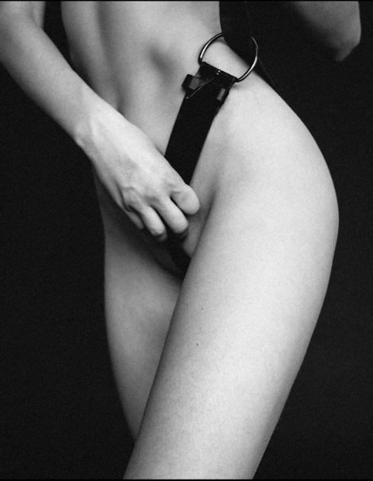 A naked white woman holding a leather belt over her private areas