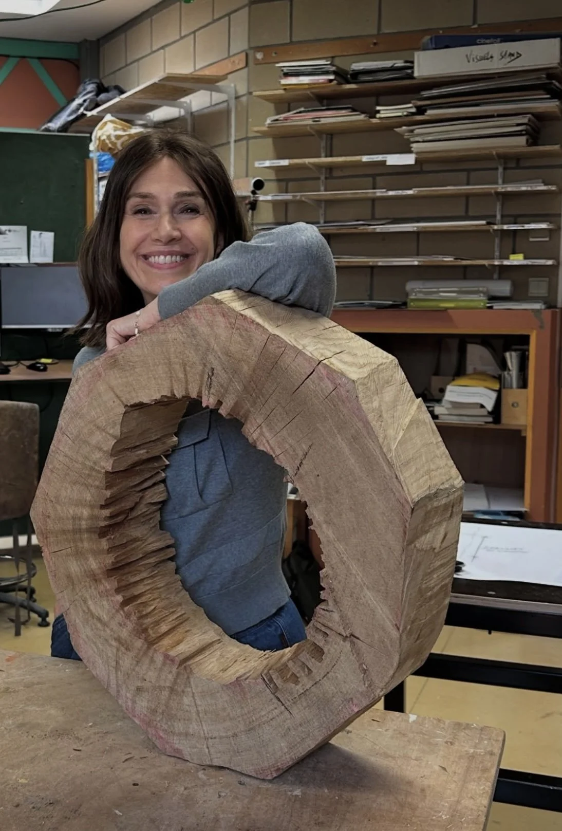 A smiling woman hugging a person with their head on her shoulder, seen through a large, roughly cut hollow wooden ring in a workshop or classroom setting.