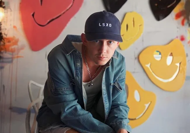 A young man wearing a blue denim jacket, white shirt, and a navy blue baseball cap with white text, sitting in front of a wall decorated with large colorful smiley face masks and graffiti art.