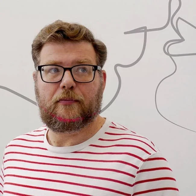 A man with glasses and a beard wearing a white shirt with red stripes, standing in front of a white wall with abstract gray line art.