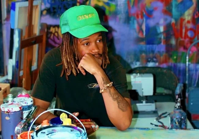 Young woman with dreadlocks wearing a green cap and black shirt, sitting at a table with art supplies and a colorful graffiti background.