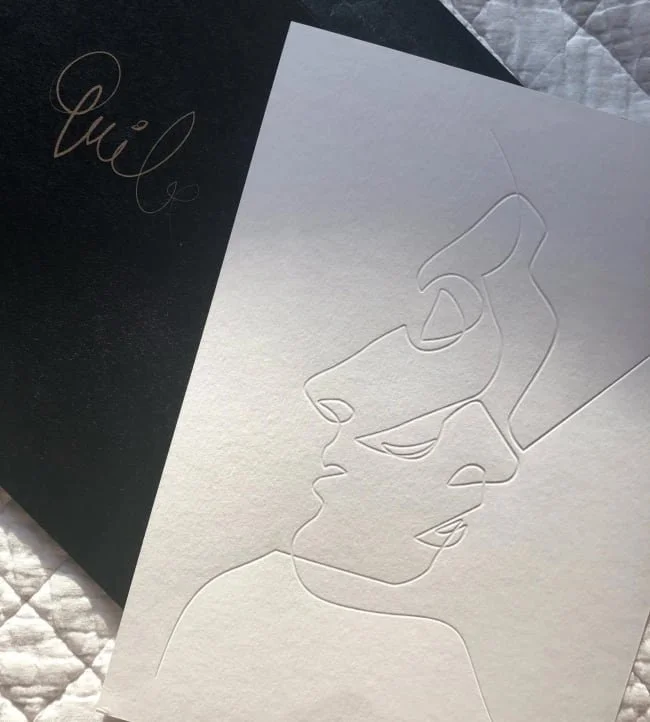 A greeting card with an embossed line drawing of a person's face in profile, partially visible with glasses, on white textured paper. The card is placed on top of a black surface with some script writing.