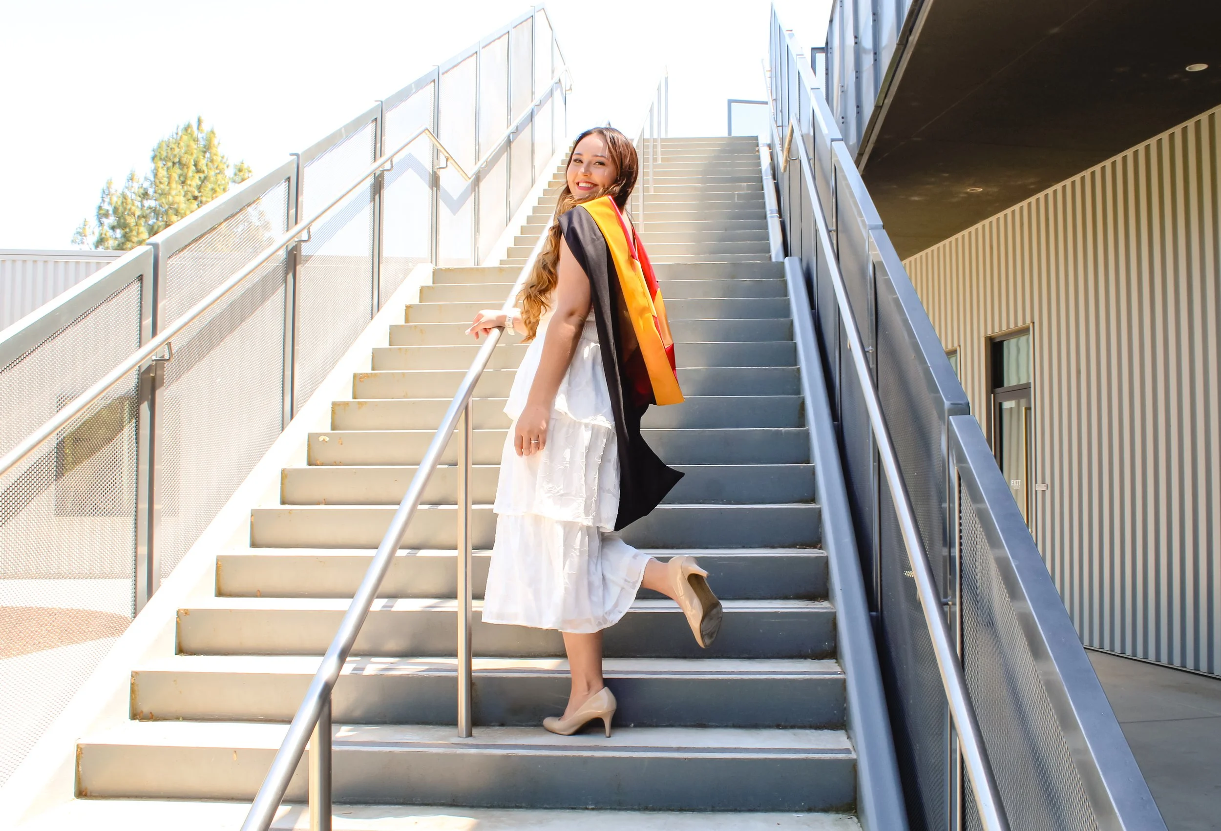 A young woman in a white dress standing on outdoor stairs, smiling, with a graduation gown draped over her shoulders, holding a backpack and wearing beige high heels.