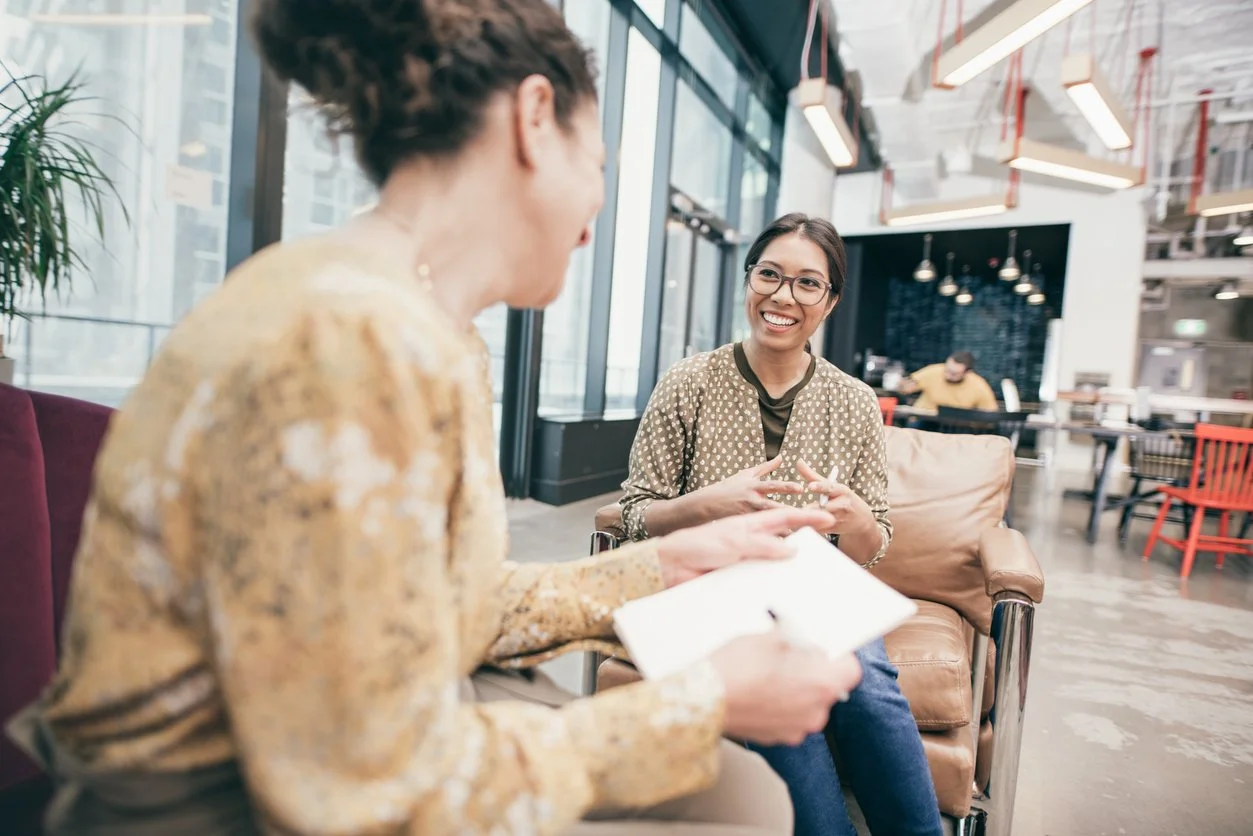 Two women having a conversation in a modern cafe, one is holding a notepad and the other is smiling, with a person working in the background.