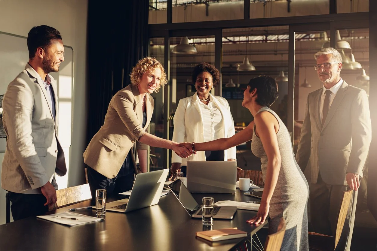 Business meeting with five diverse professionals, two women shaking hands, laptops, notebooks, glasses of water on the table in a modern office.