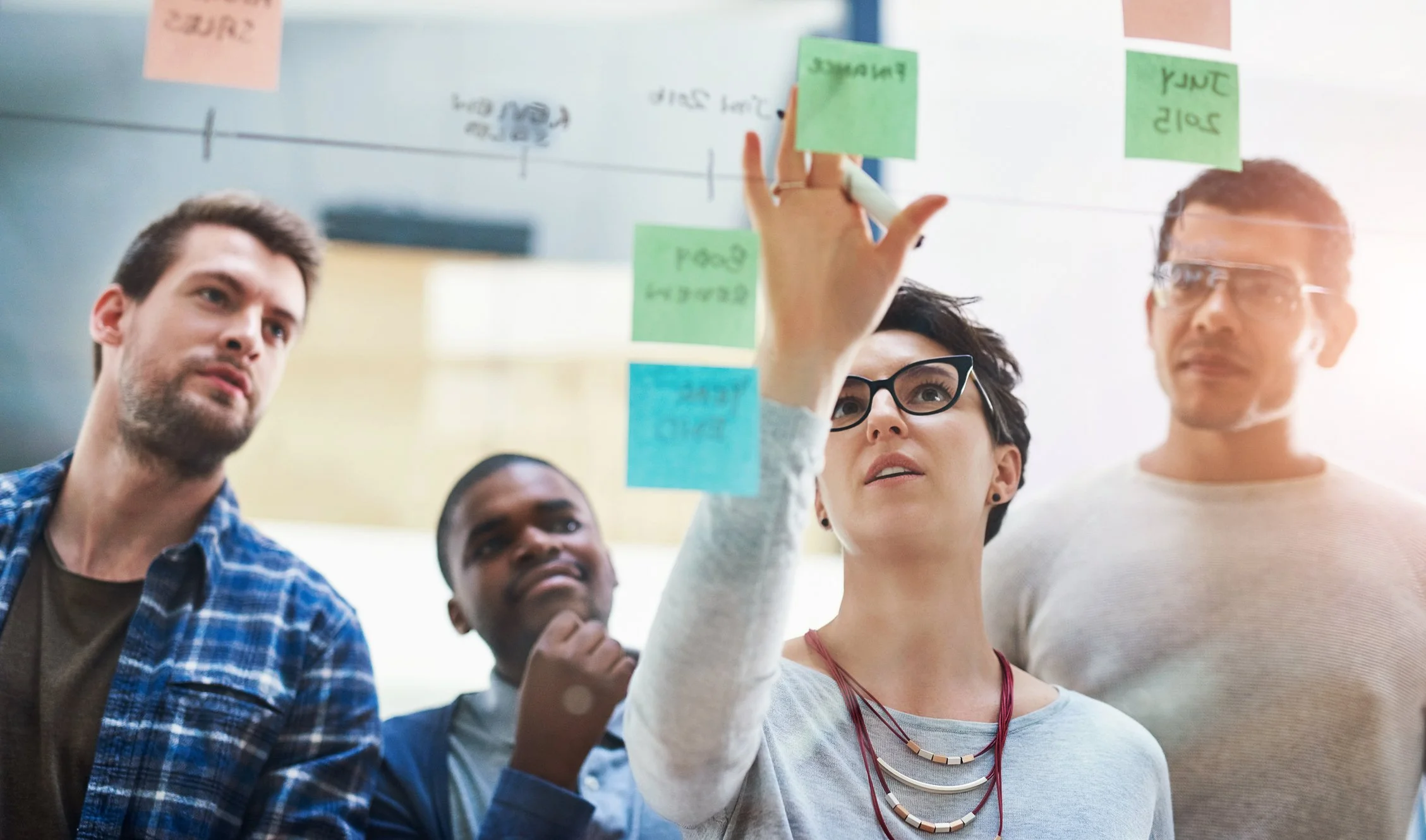 A diverse group of four people in a meeting room analyzing a timeline with sticky notes on a glass board.