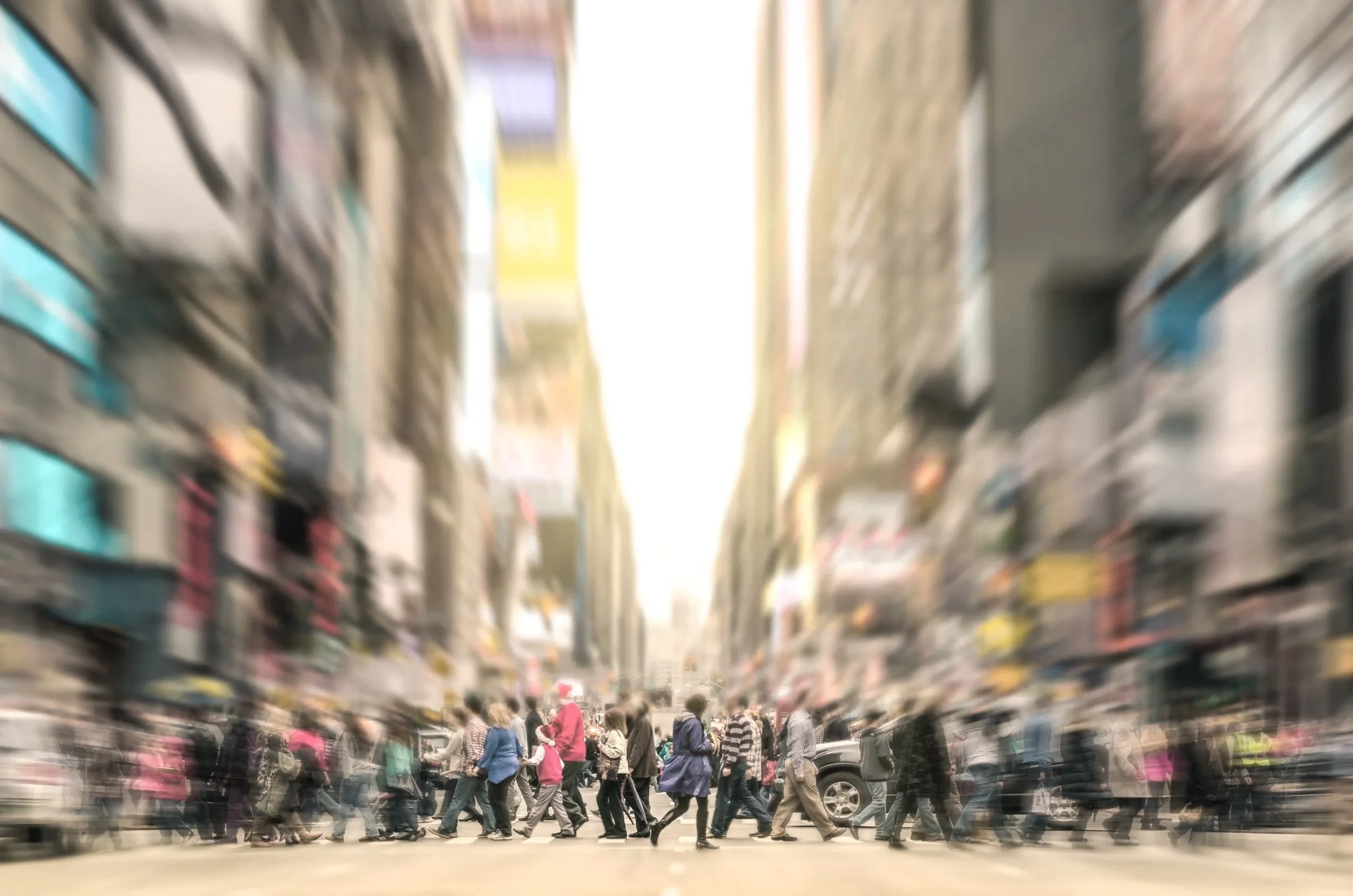 Blurred photo of a busy city street with many pedestrians crossing intersection, tall buildings on both sides, and a bright sky overhead.