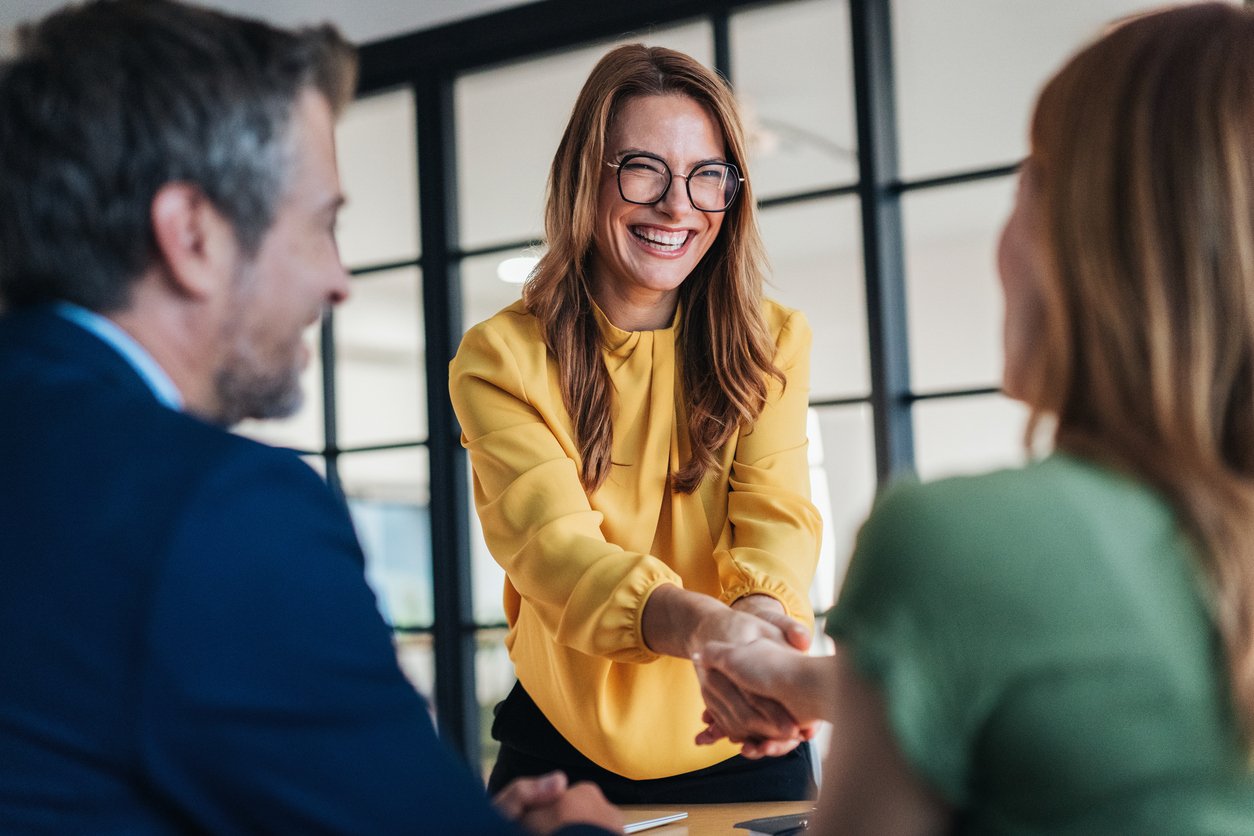 A woman with glasses in a yellow blouse smiling and shaking hands with a woman in green, while a man in a blue suit watches in an office setting.