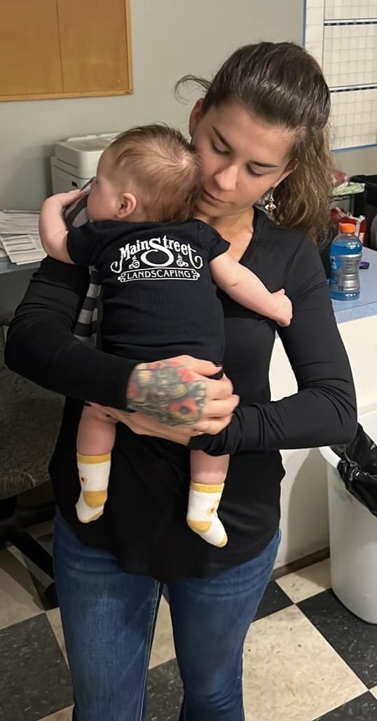 Main Street Landscaping team member and daughter of owner holding baby wearing branded onesie, representing the family-run, multi-generational business
