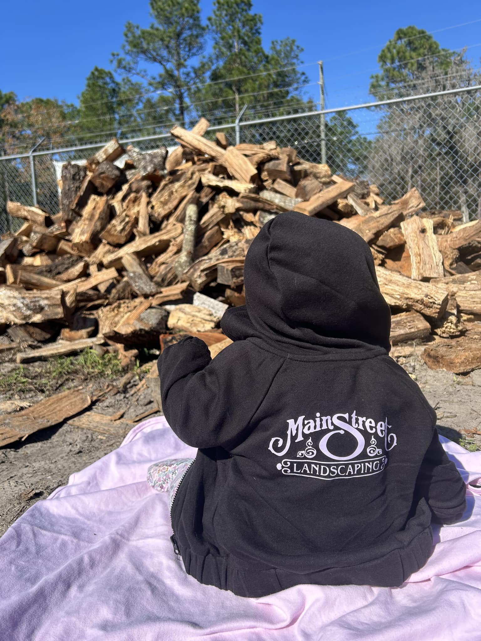Child wearing Main Street Landscaping hoodie in front of stacked firewood, symbolizing family legacy and seasonal service