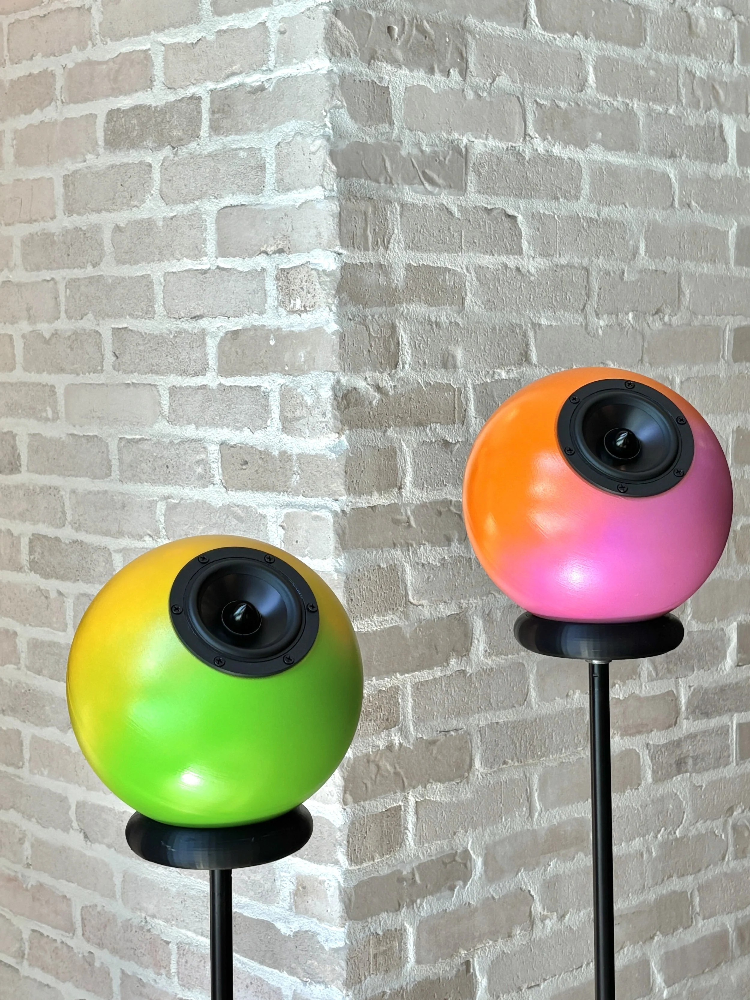 Colorful spherical speakers, one green and yellow, the other pink, are mounted on black stands against a white brick wall.