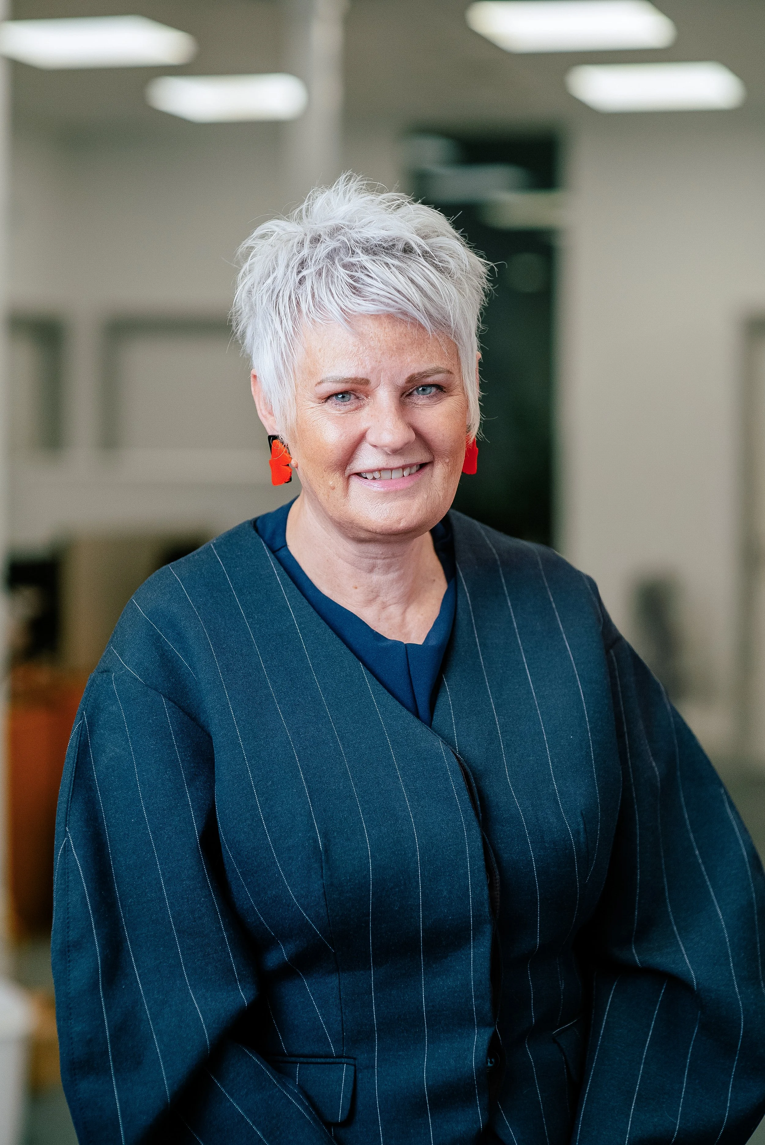 A smiling woman with short, spiky white hair, wearing a dark blue pinstripe blazer and red earrings, standing in an office environment.