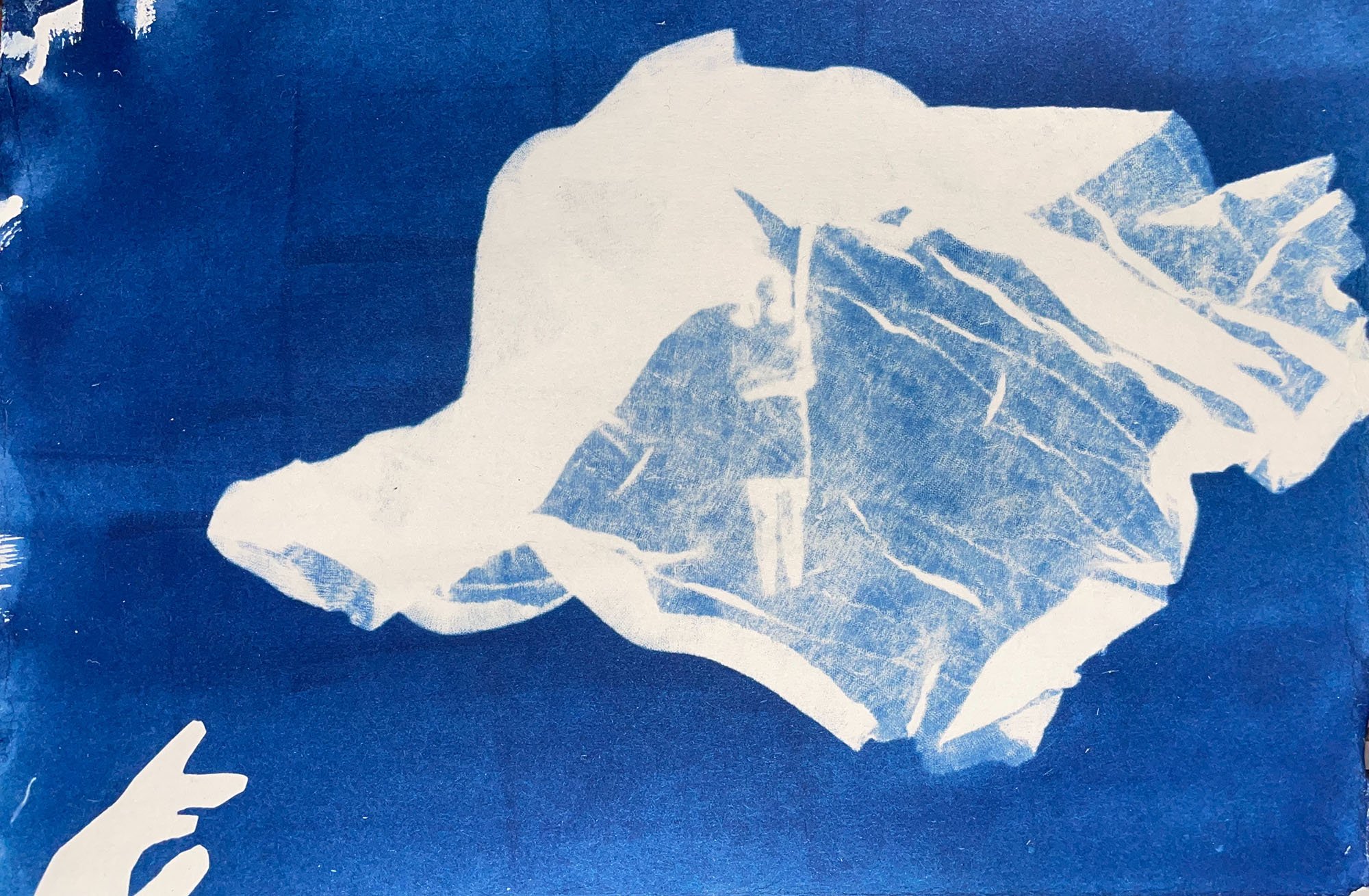 A deep blue cyanotype print with chiffon obscuring an image of a woman, in the bottom lefthand corner there is a hand reaching up towards the woman