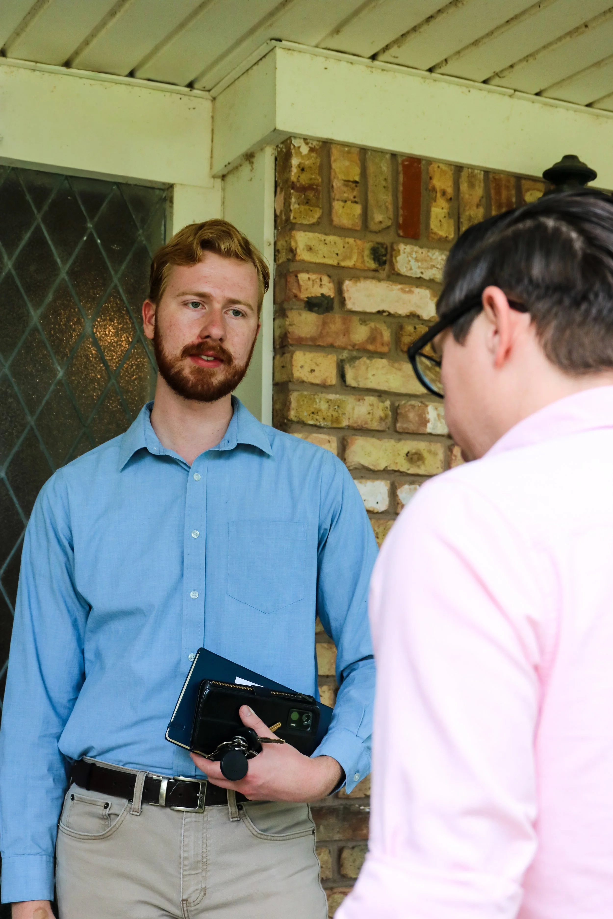 Two men having a conversation outside a building with brick wall and diamond-patterned window.