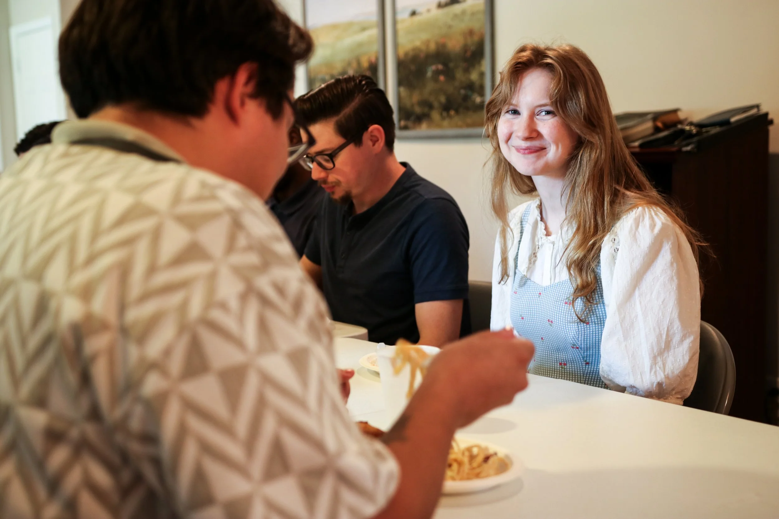 A young woman with long red hair and a white blouse smiling at the camera while sitting at a table with a man in glasses and a dark shirt, and other people, in a room with framed artwork on the wall.