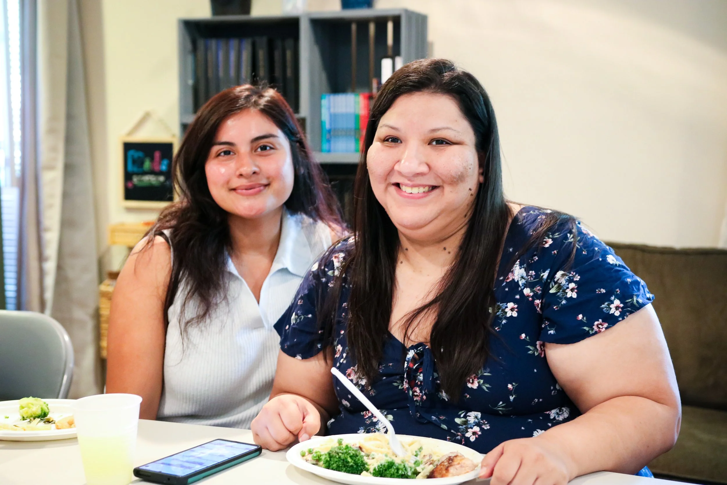 Two women sitting at a table with plates of food, smiling at the camera in a cozy indoor setting with bookshelves in the background.
