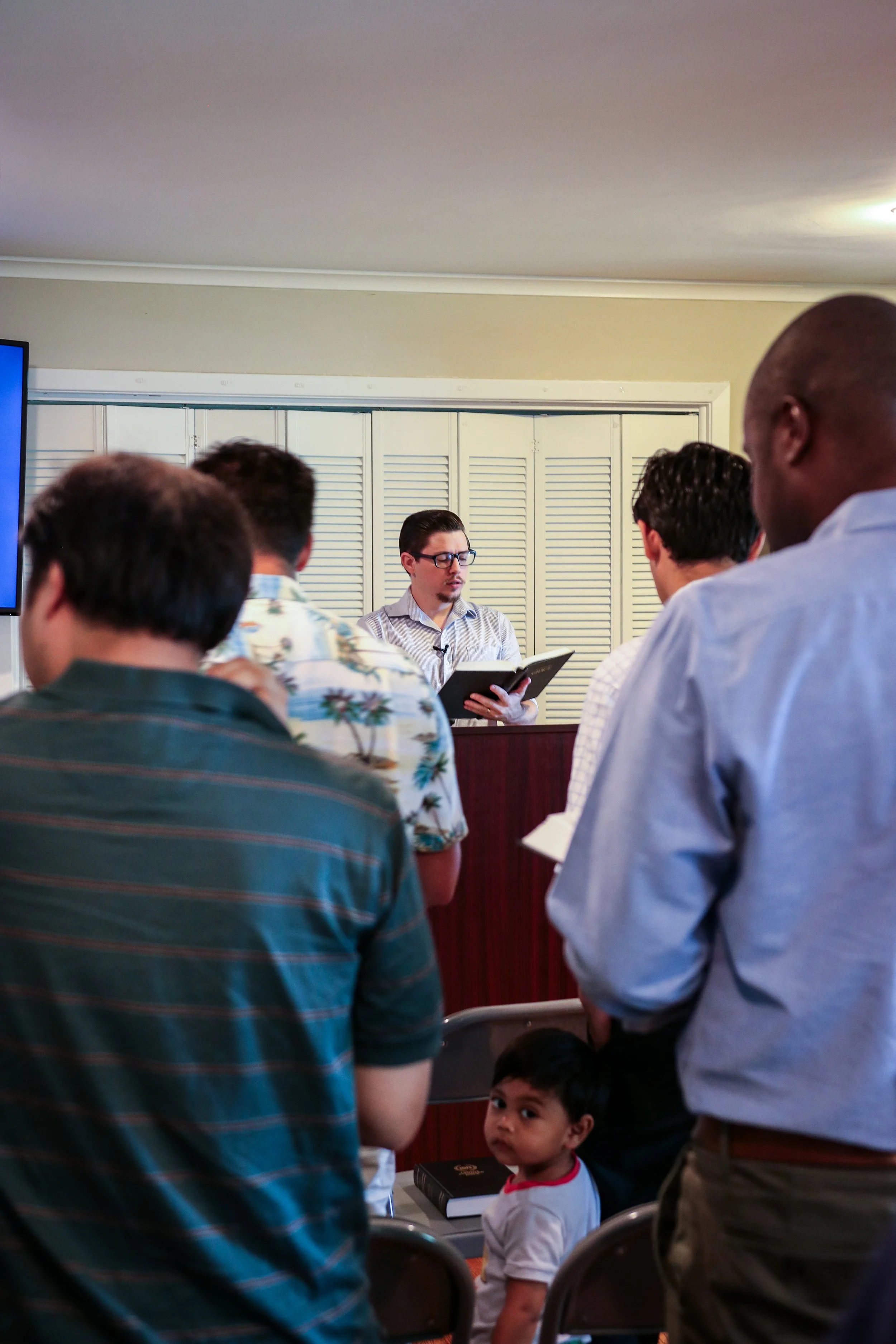 A man with glasses and a striped shirt reading from a book at a podium during a church service, surrounded by congregants.