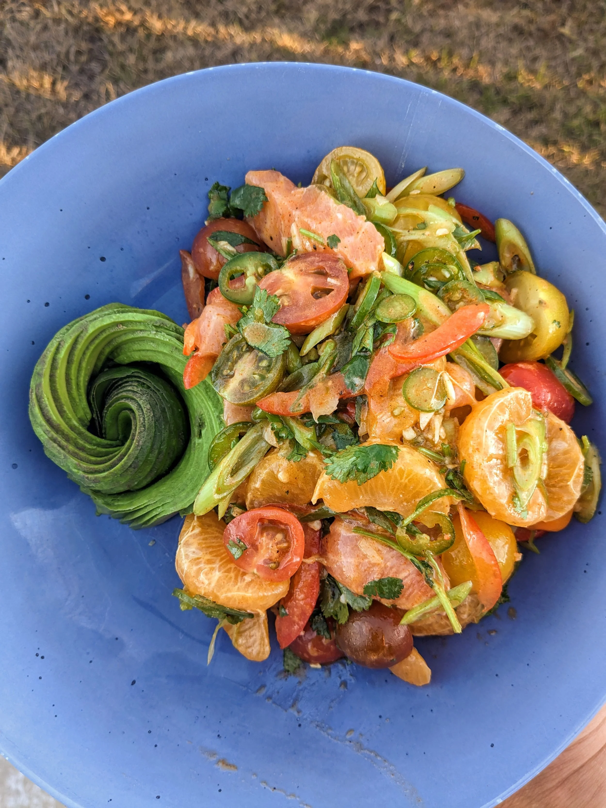 A blue bowl of fresh-mixed salmon poke salad with orange slices, cherry tomatoes, sliced green onions, cilantro, and chili peppers, garnished with an avocado rose on the side.