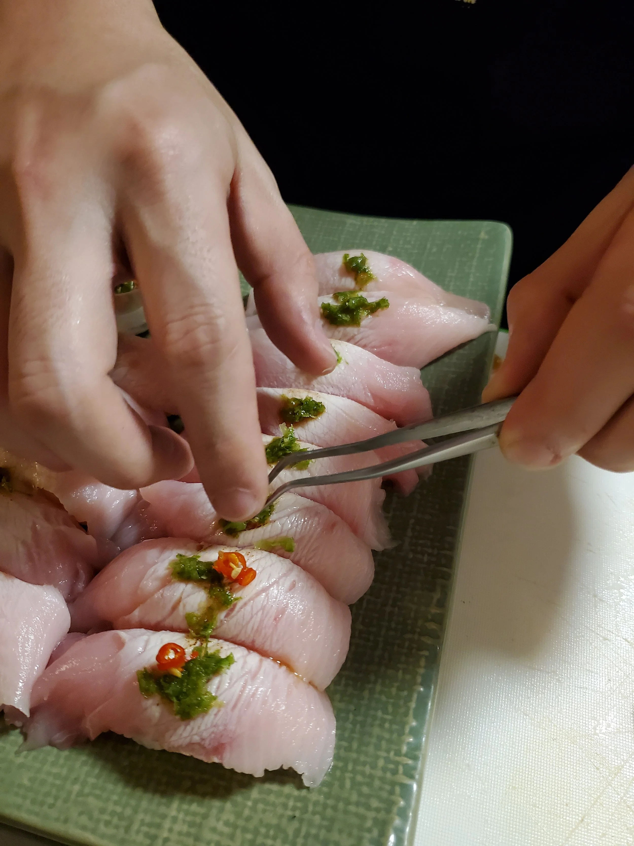 Close-up of hands preparing raw fish sashimi on a green plate, garnished with green herbs and red chili slices.