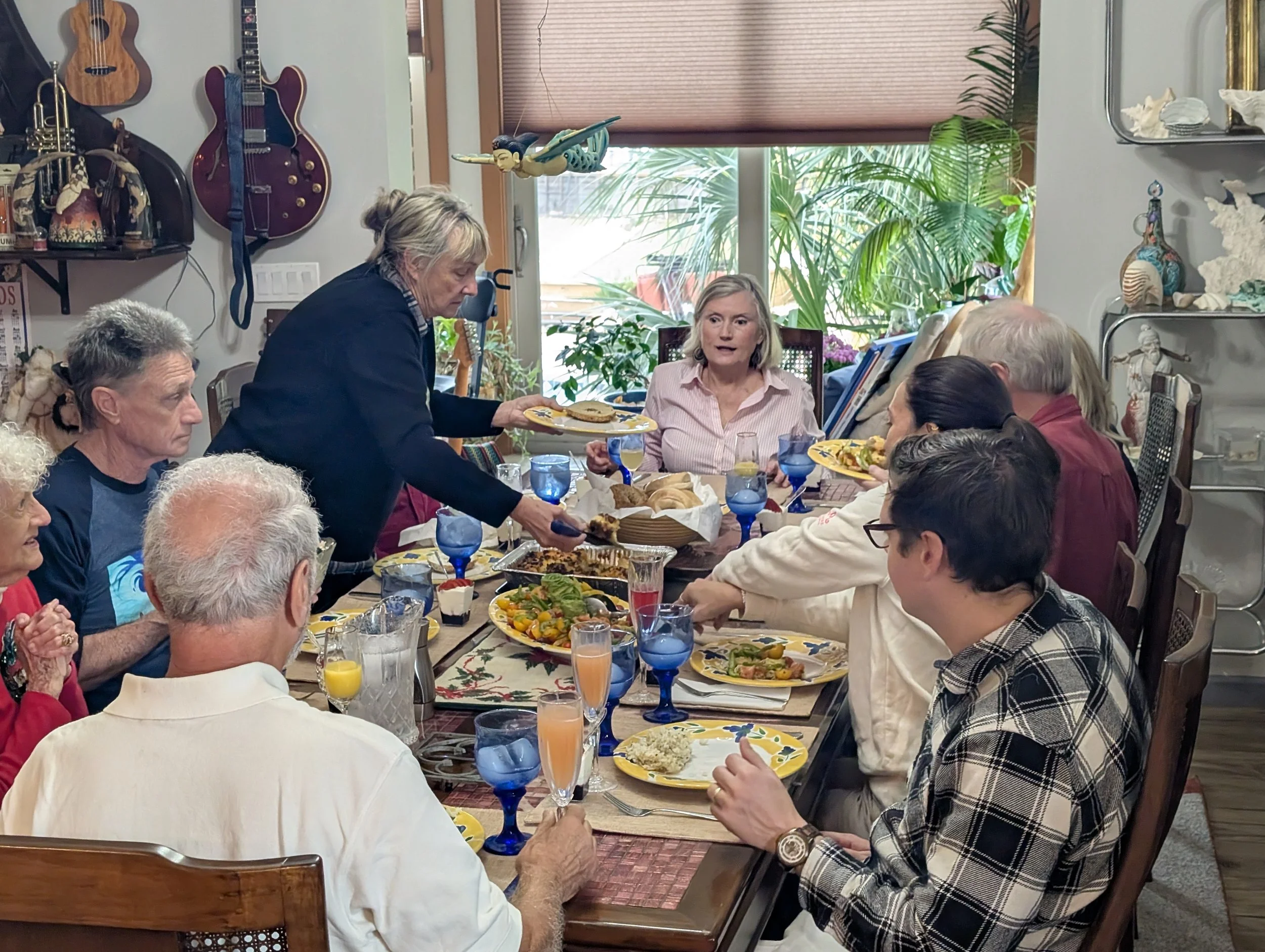 People gathered around a dining table enjoying a meal, with a woman serving food. The table is set with various dishes, colorful plates, and drinking glasses. The background shows a window with plants and decorative items on shelves.