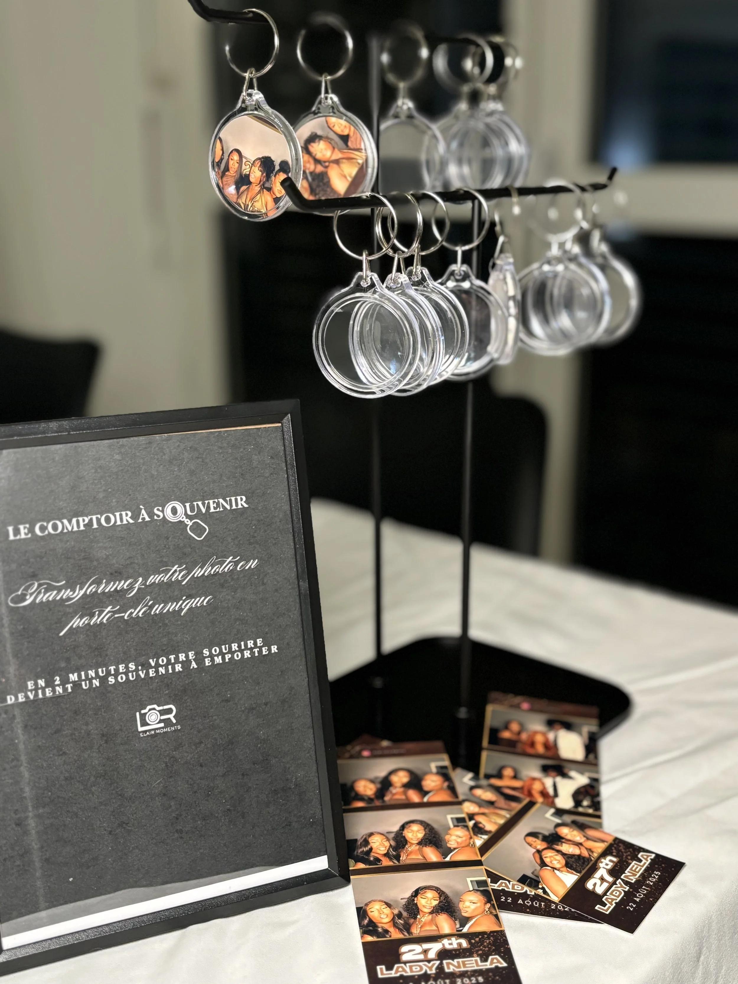Un présentoir à porte-clés avec des photos imprimées, accompagné d'une affiche invitant à transformer une photo en souvenir à emporter, et des coupons pour un événement célébrant Lady Nela, le 22 août 2025.