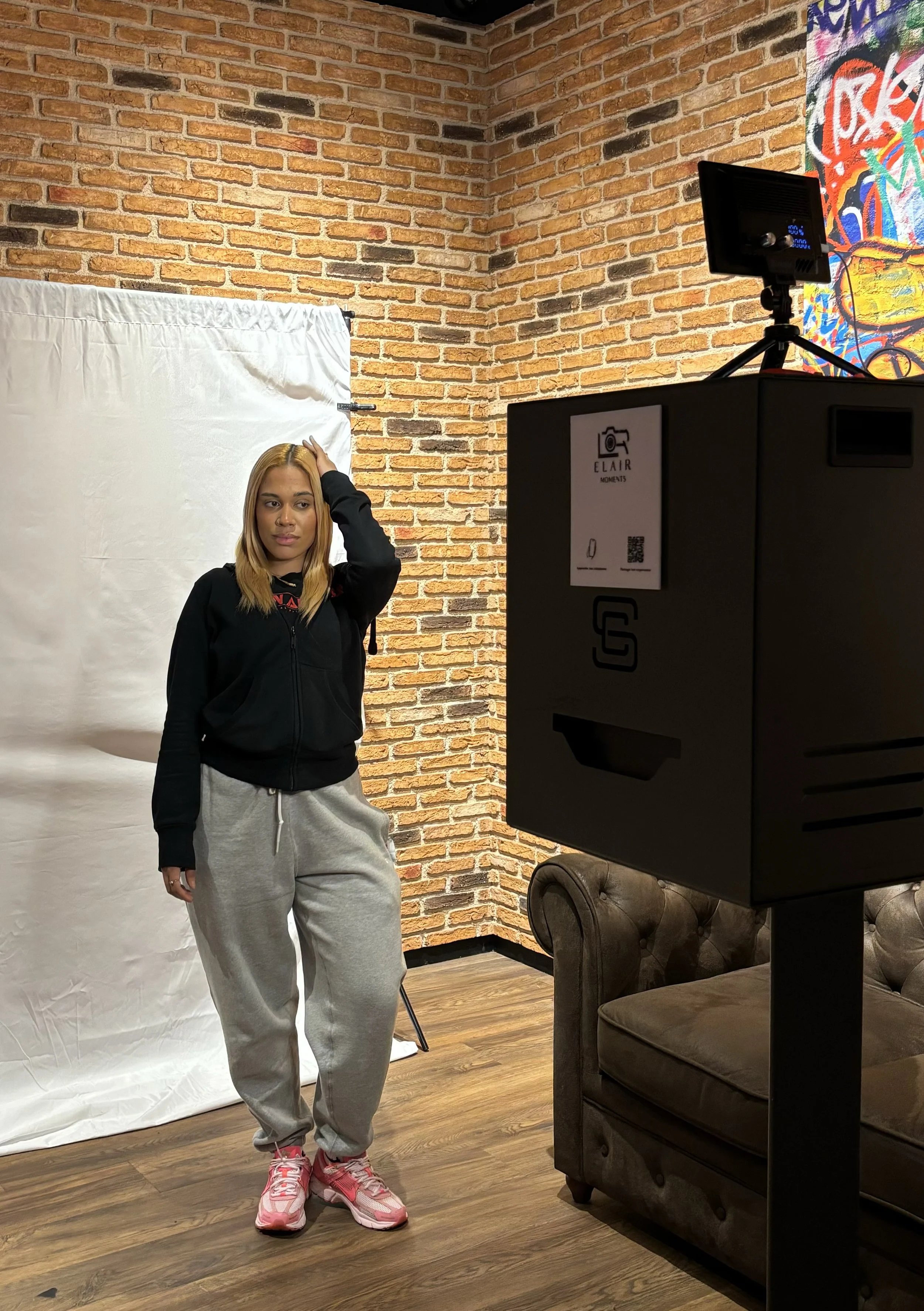 Une femme pose devant un fond blanc dans un studio, avec un mur en briques en arrière-plan, et un grand appareil photo noir sur un support proche.