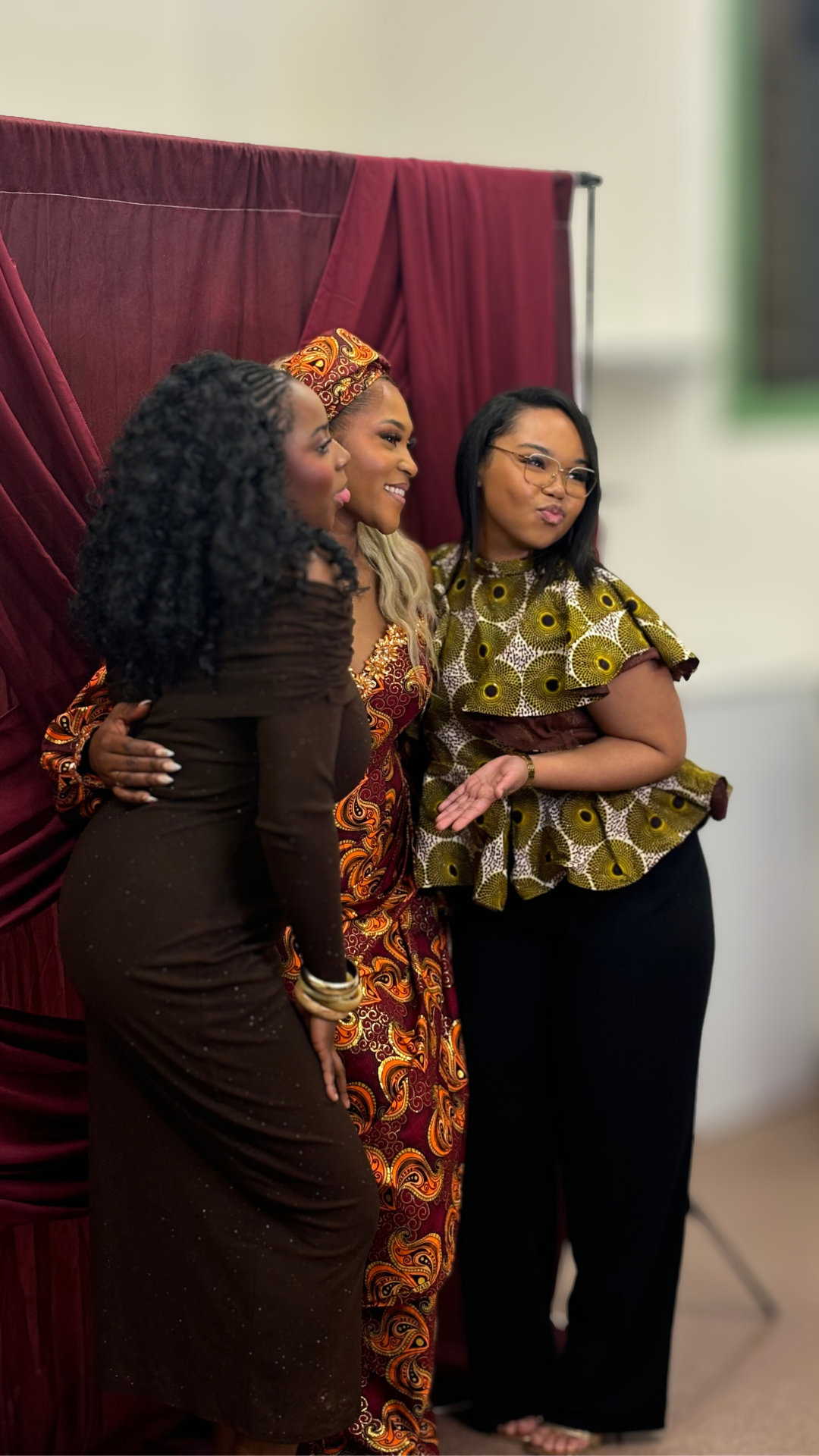 Trois femmes posant et souriant pour une photo, devant un panneau de tissu bordeaux, portant des vêtements africains colorés et élégants.
