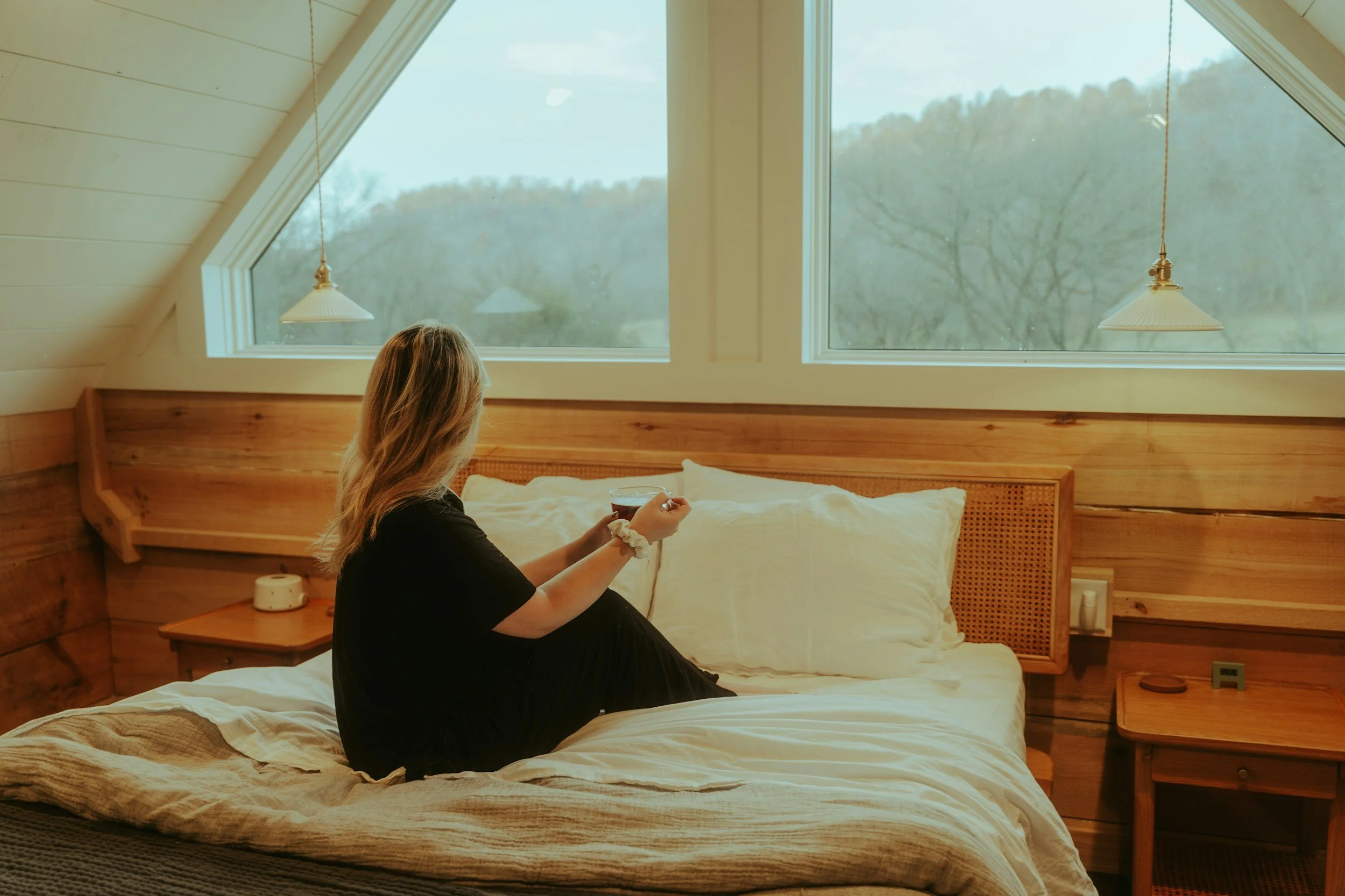 Woman peacefully sitting on her bed with a cup of coffee looking out her window to trees, representing trauma-informed therapy safety and nervous system regulation after a therapy intensive. Camas, WA