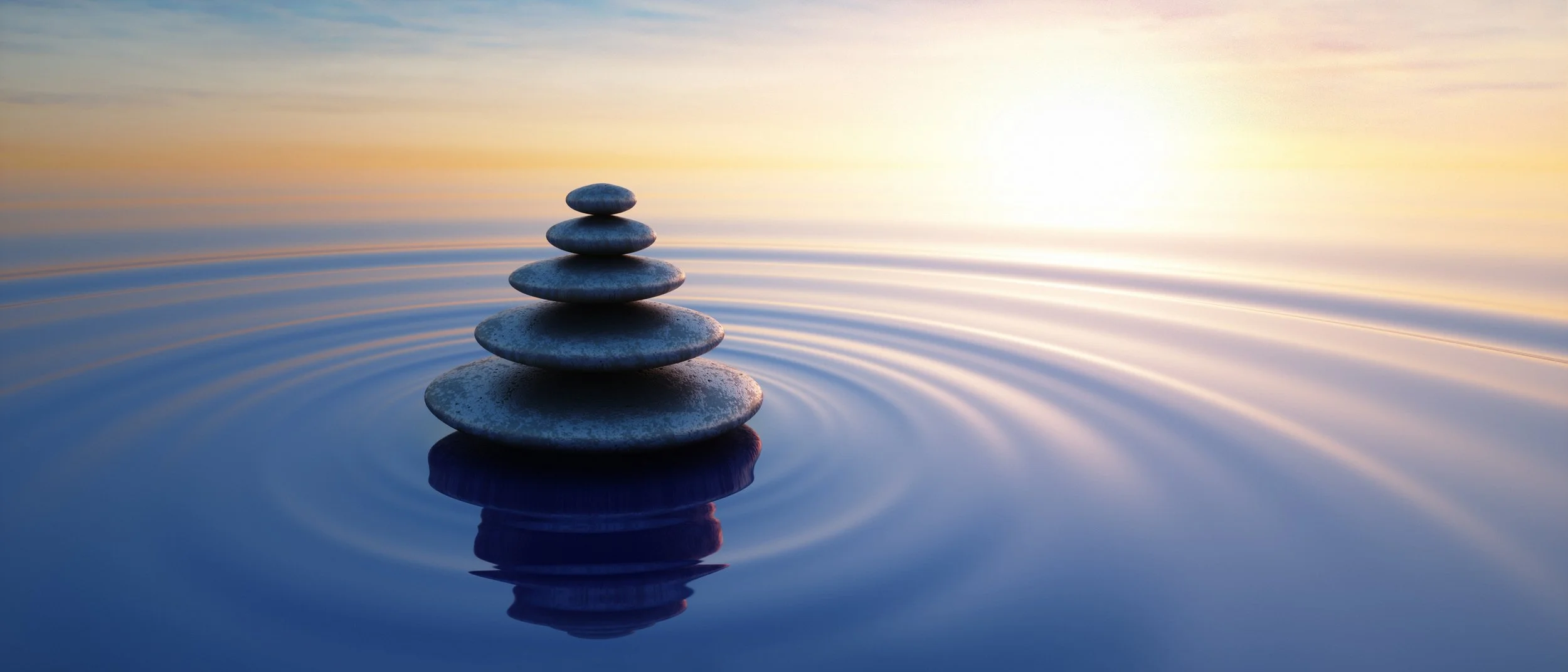 Stacked smooth stones balancing on one another on a calm water surface with ripples, during a serene sunset or sunrise.