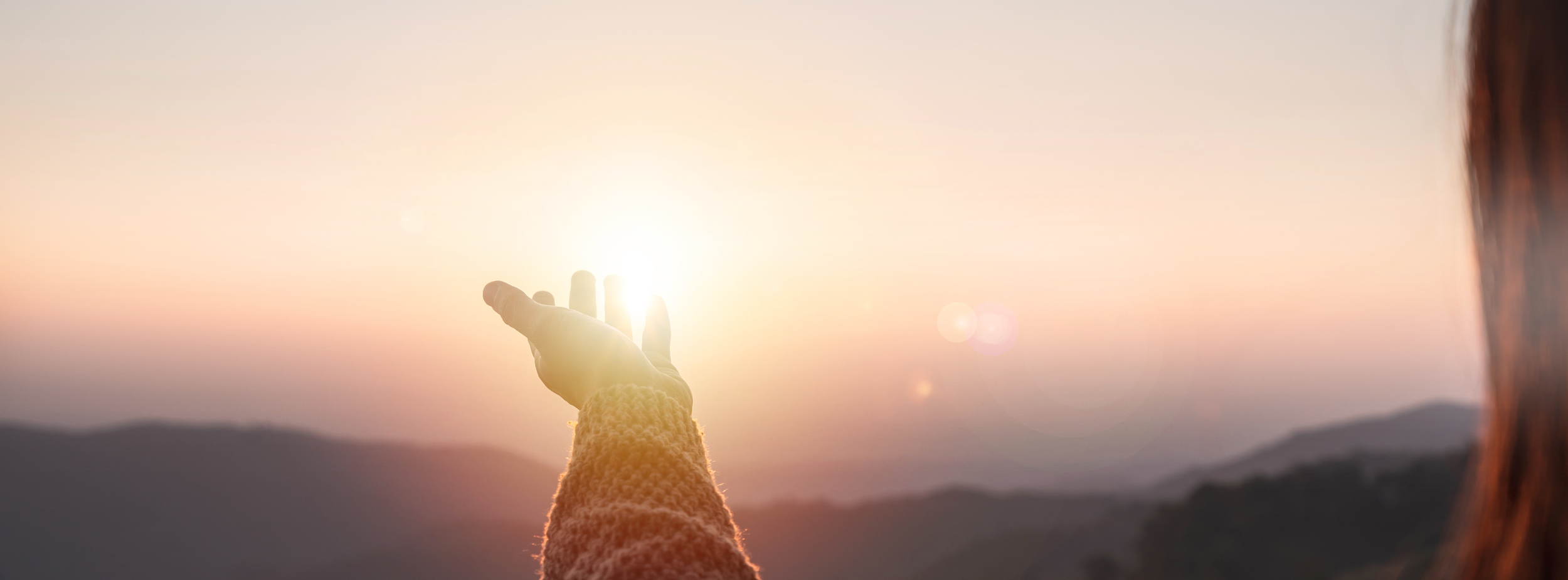 Person reaching out towards the sun during sunset or sunrise, with mountains in the background.