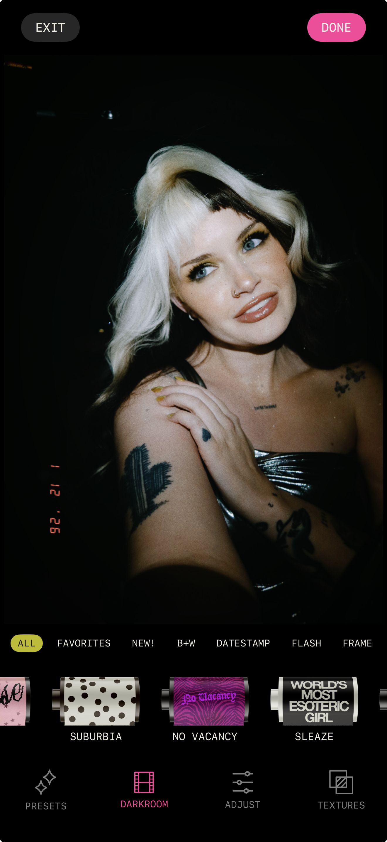 A woman with black and platinum blonde hair, blue eyes, and tattoos on her arm and chest, posing for a photo with her hand on her shoulder, in a dark setting.