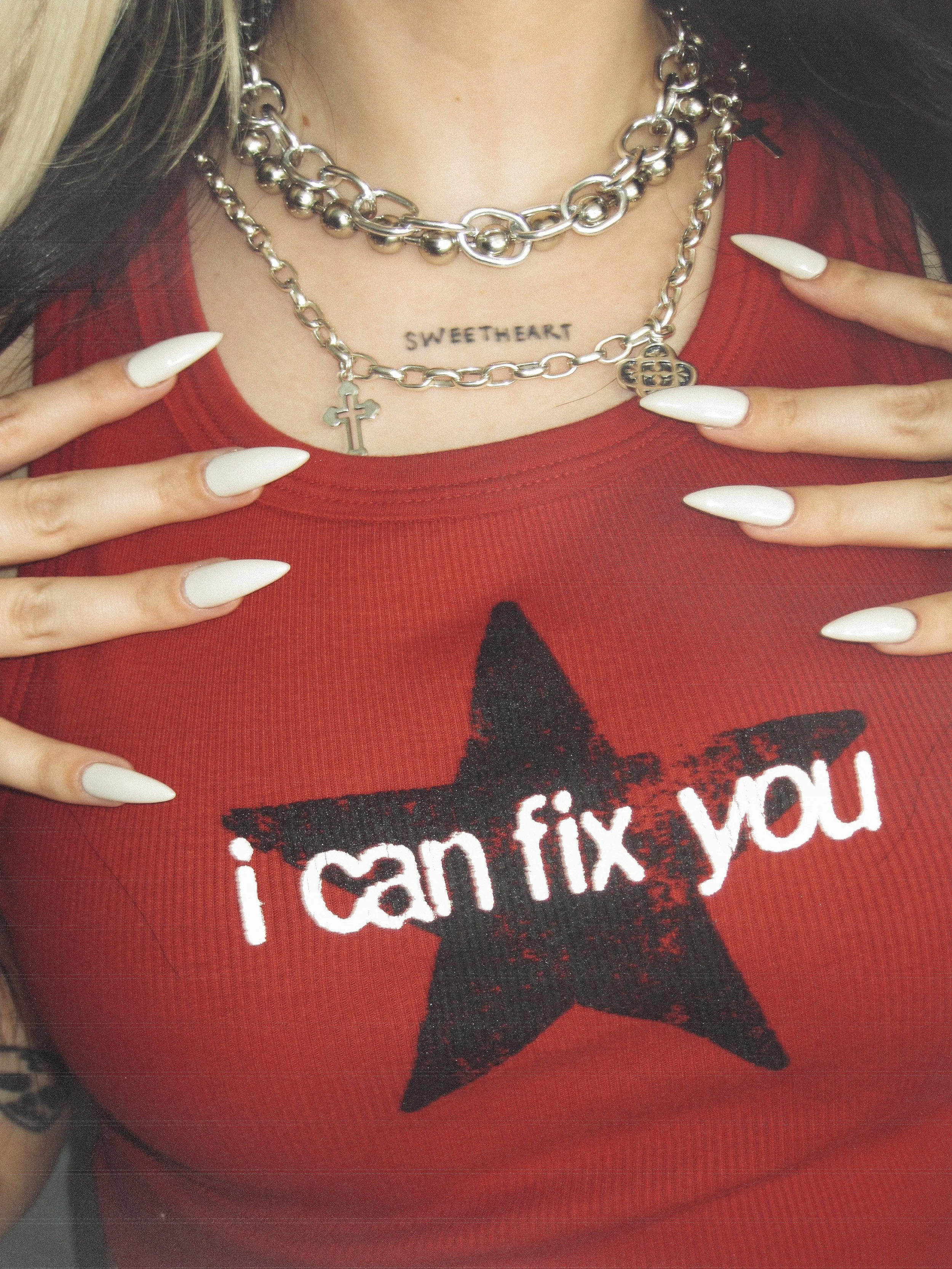Person wearing layered silver necklaces, a red shirt with a black star design and the words 'i can fix you' printed on it, and showing long white-painted nails.
