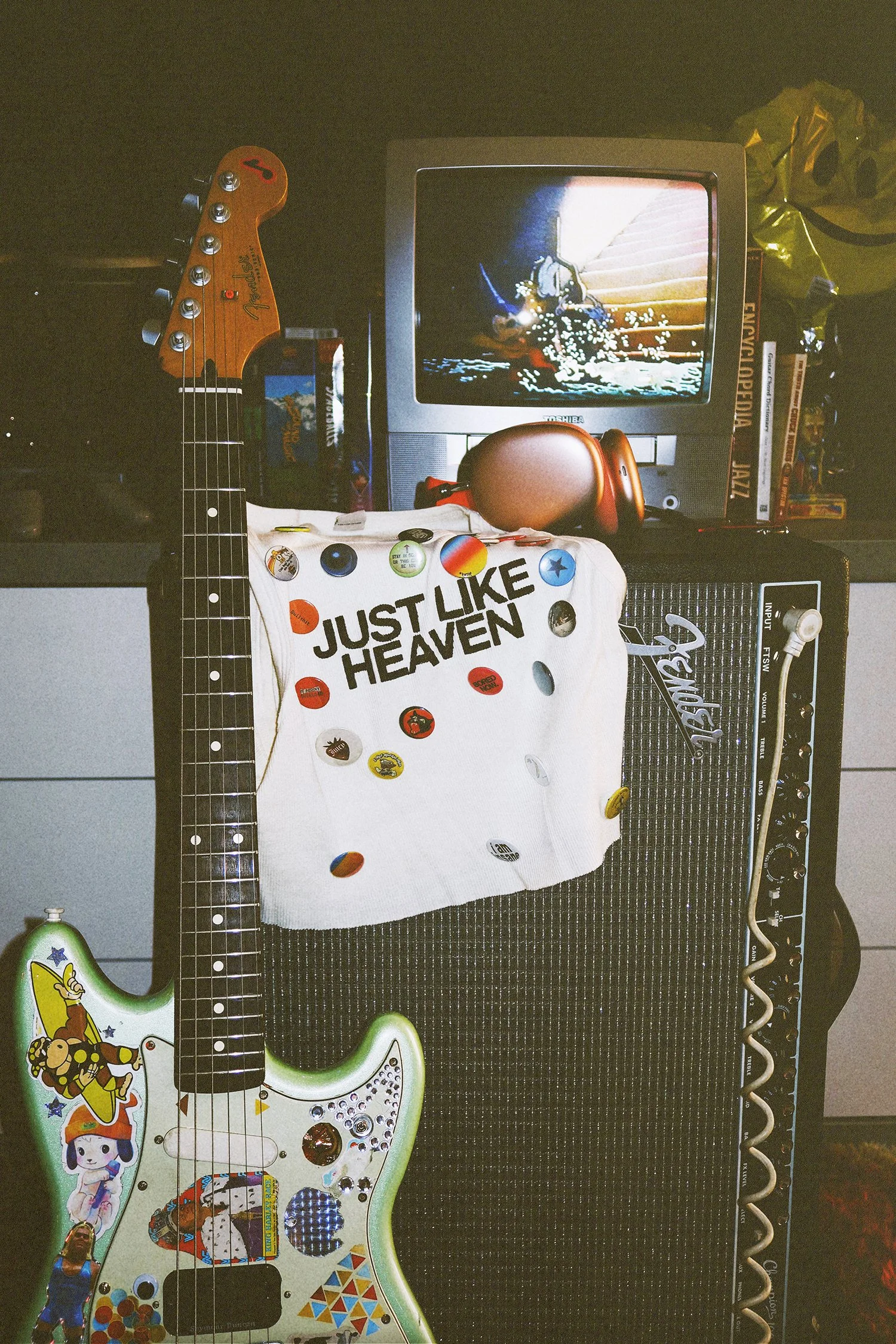 A collection of musical items including a decorated guitar, a vintage television displaying an animated scene, a white cloth with the text 'JUST LIKE HEAVEN' and scattered buttons, a Fender amplifier, and various books and objects in the background.
