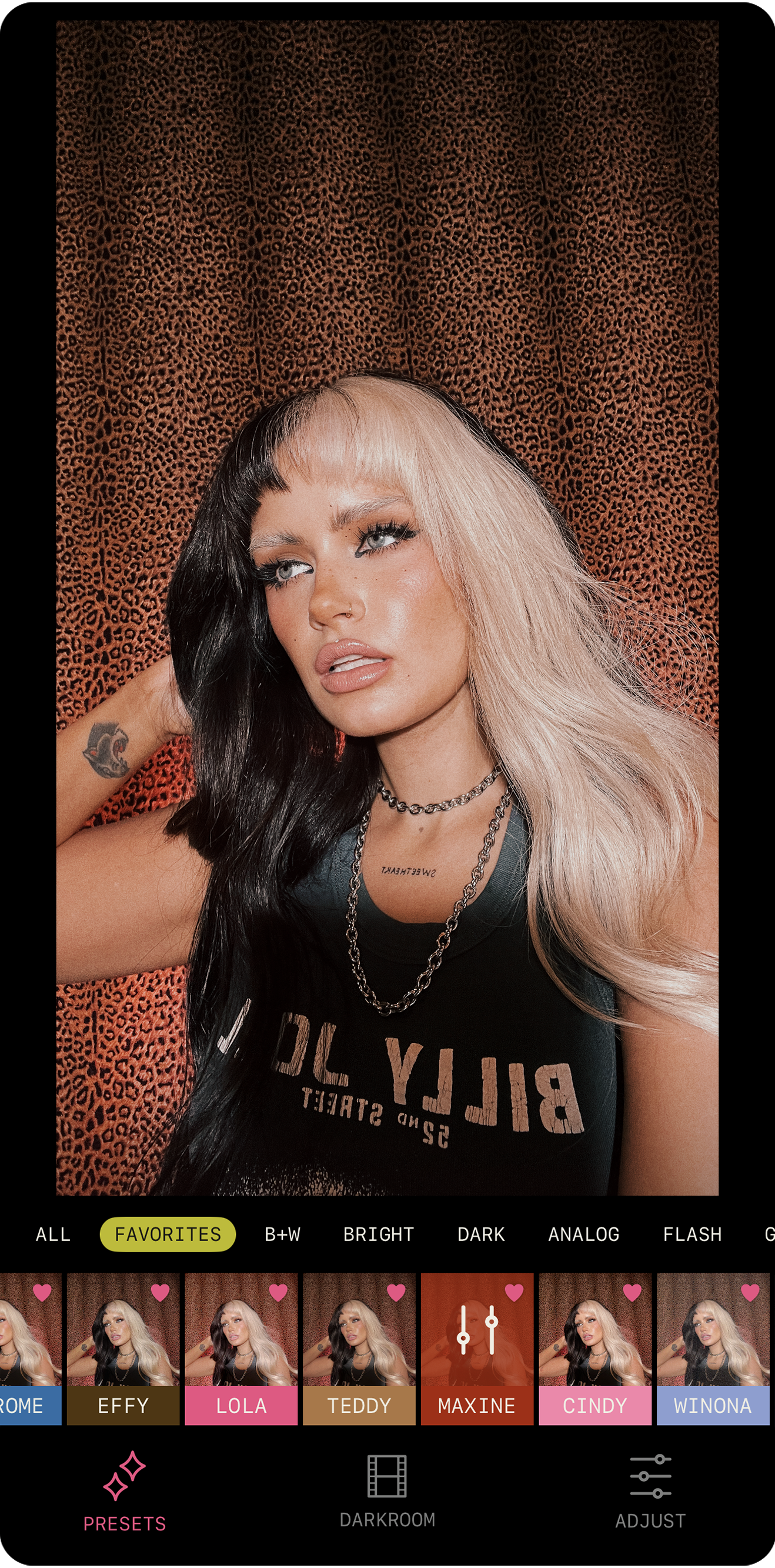 Young woman with black and blonde hair, tattoos, wearing a black t-shirt, jewelry, in front of a leopard print background.