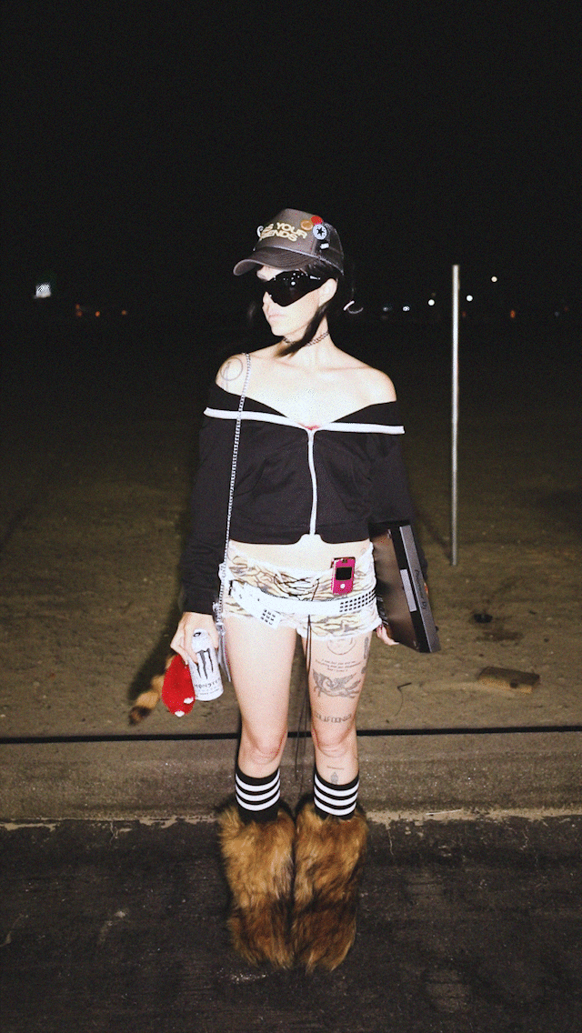 A person dressed in a black off-shoulder top, tiger-print shorts, striped knee-high socks, and furry animal slippers standing outdoors at night, holding a can and a laptop, wearing sunglasses and a cap.