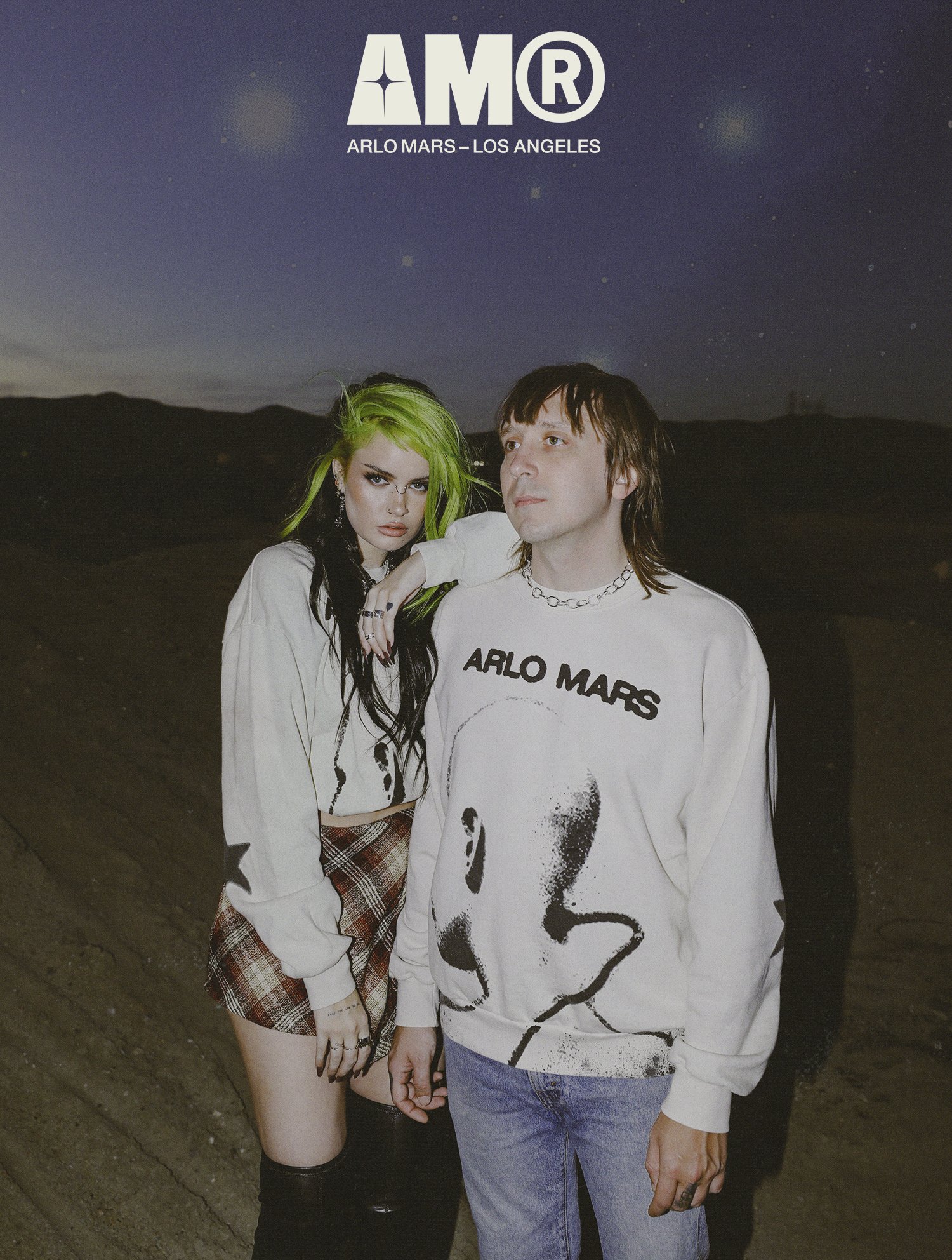 Two people standing outdoors at night, a woman with green and black hair wearing a white jacket and plaid skirt, and a man with brown hair wearing a white sweatshirt with a face graphic and the words "ARLO MARS," in a desert landscape.