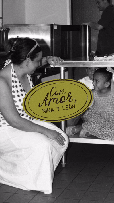 A woman and two young girls sitting on the ground, with the woman leaning forward and engaging with the girls. There is a sign with the words "Con Amor, Nina y León" positioned in front of the women and girls, indicating a gesture of love from Nina and León.