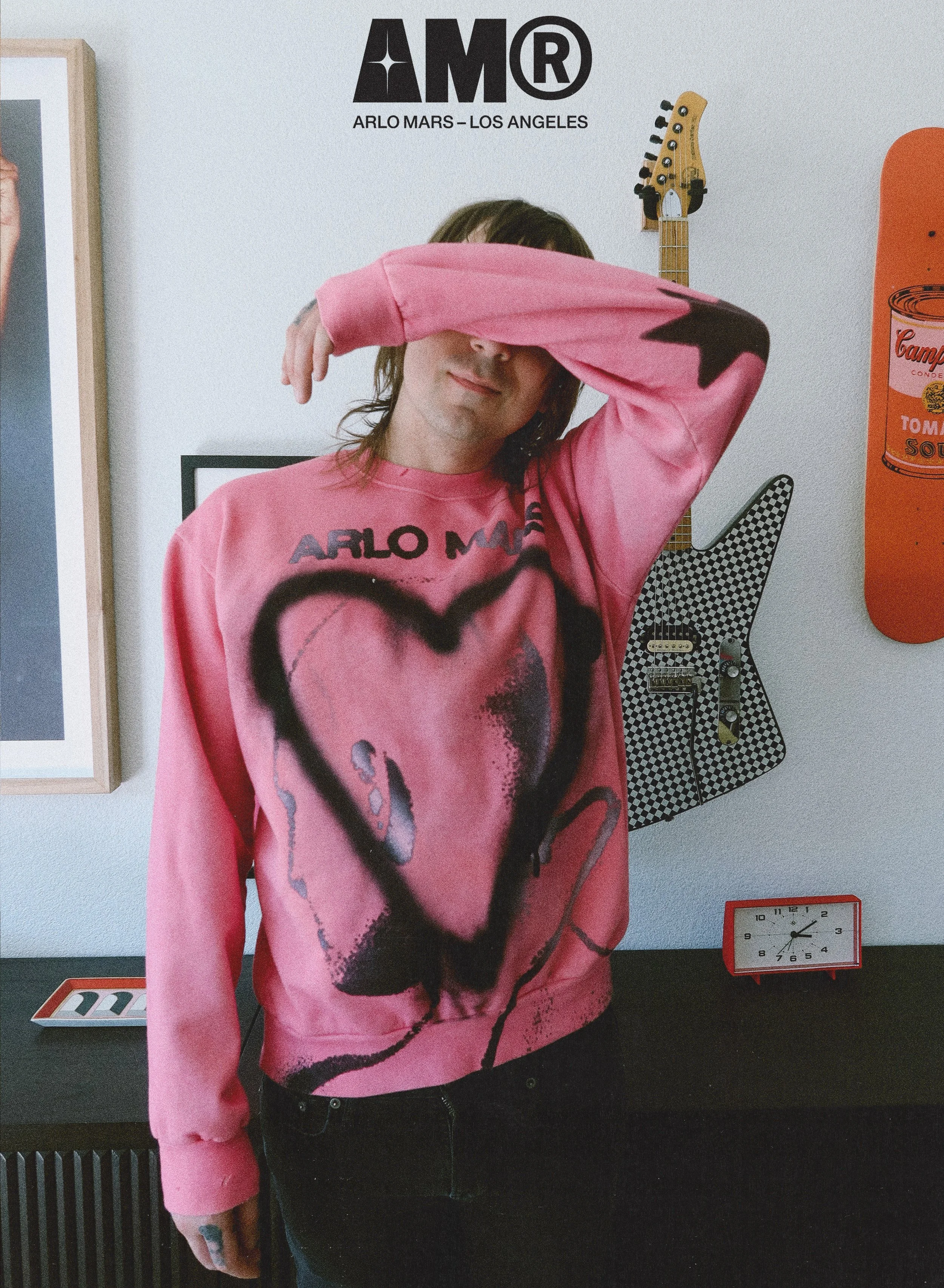 A man in a pink sweatshirt with graffiti-style artwork and the words 'ARLO MARS' stands with his arm across his face inside a room decorated with music and pop art. A guitar and skateboard are on the wall, a small red clock on a black table, and framed artwork are visible in the background.