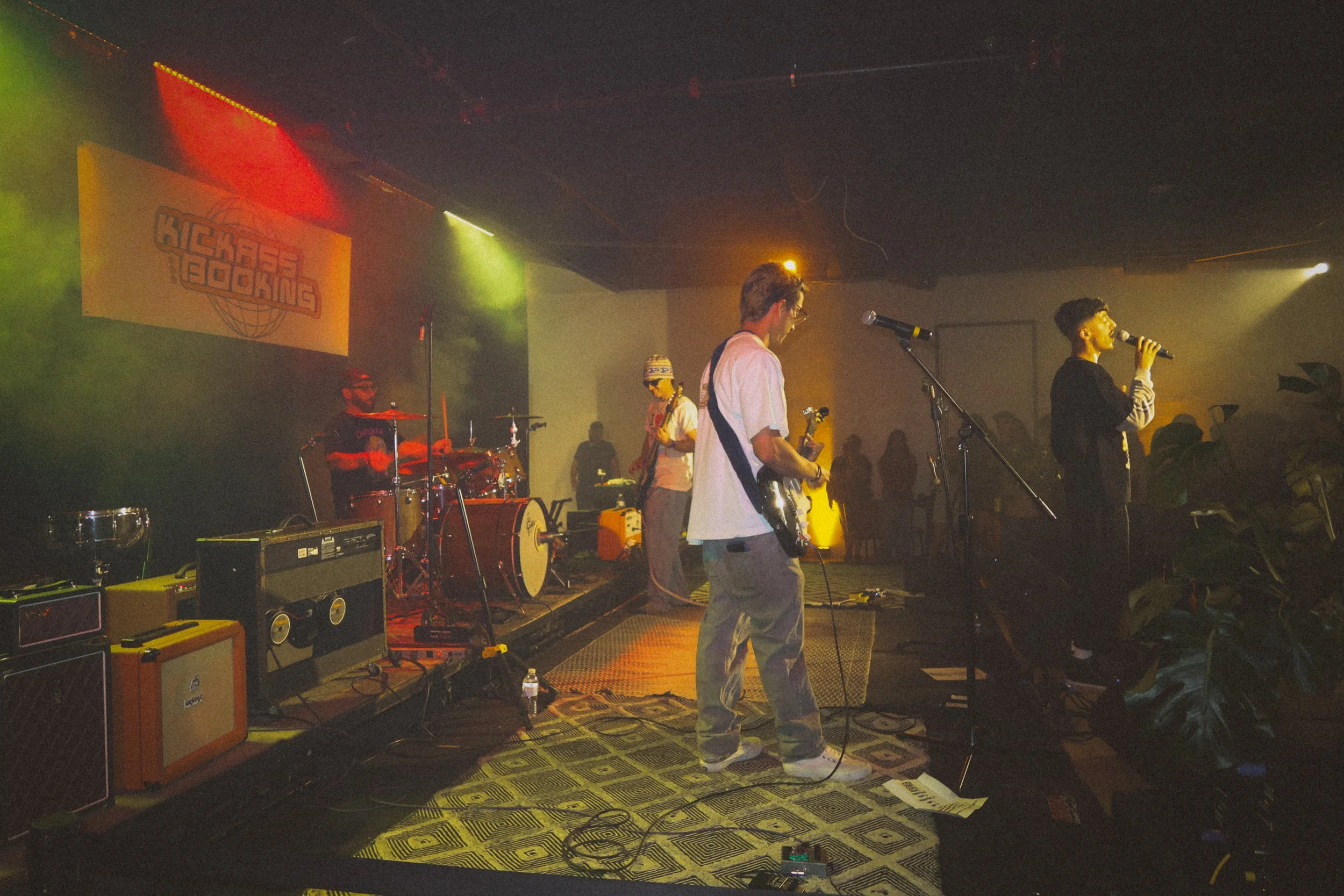 Band performing on stage with colorful lighting and a large screen showing a logo in the background, audience visible in silhouette.