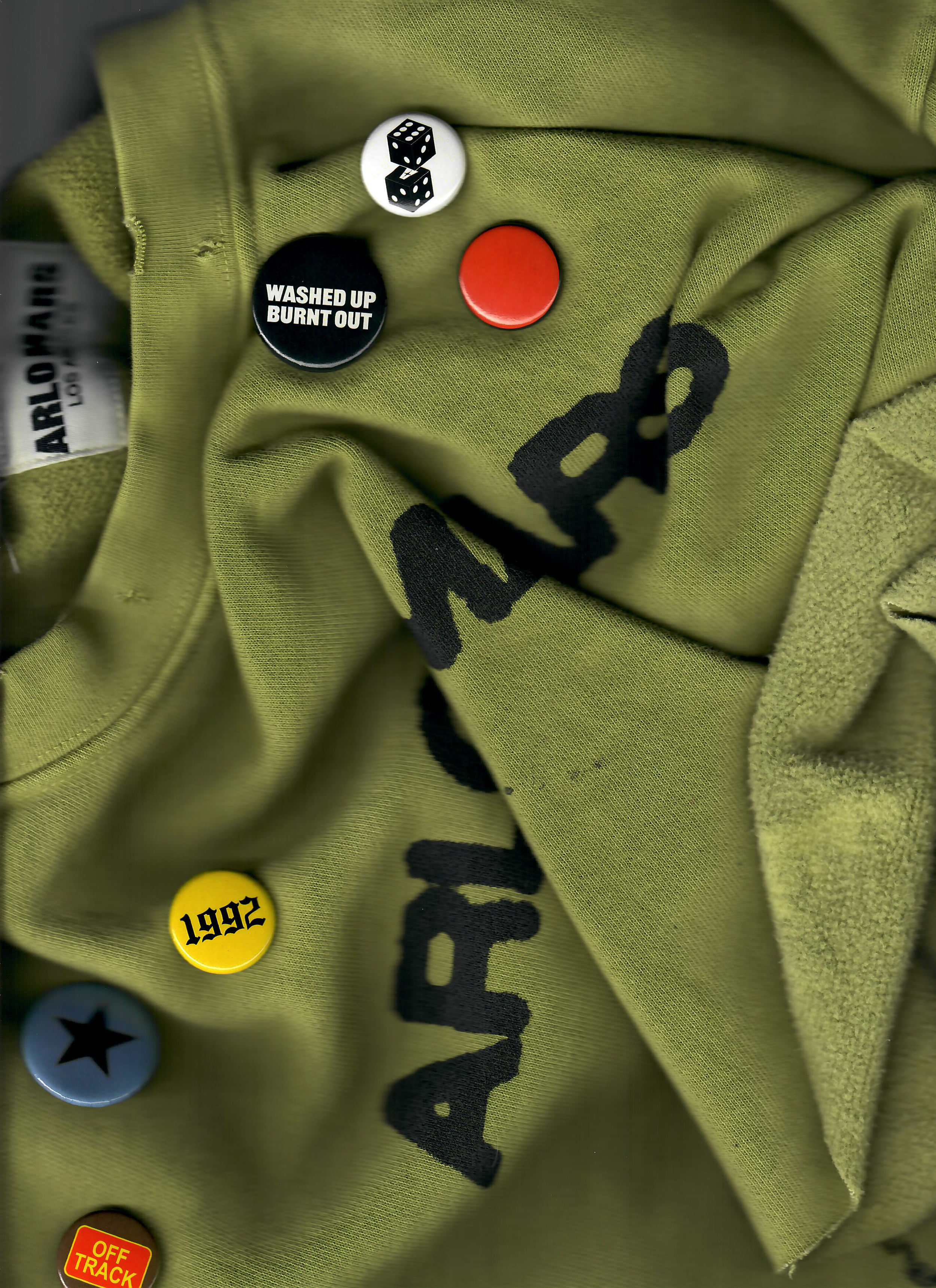 Close-up of a green fabric with black printed text, showing several colorful buttons and pins, including a black pin with white text 'WASHED UP BURN OUT', a yellow pin with black text '1992', a blue pin with a black star, a red pin, and a black and white dice pin. Part of the ARBOR brand tag is visible on the left side.