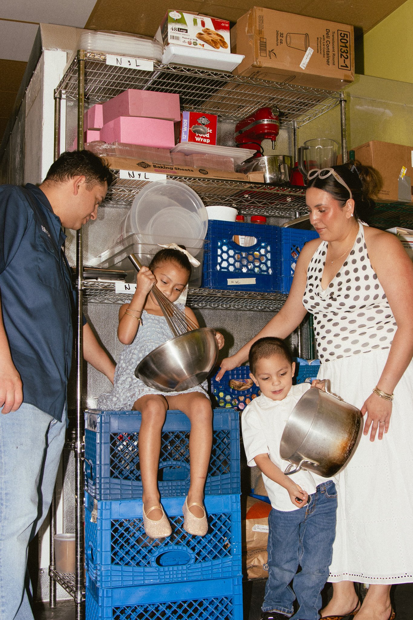 A family of four is in a storage room, engaging in a playful activity with kitchen pots and a mixing bowl. The man and woman are supervising their two children, who are holding and using kitchen utensils.