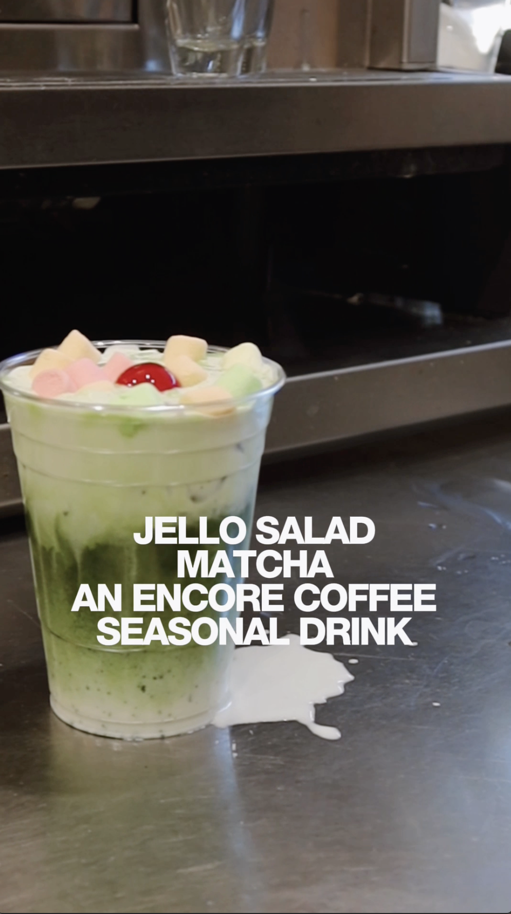 A clear plastic cup filled with a layered green matcha drink topped with marshmallows and a cherry. The cup is placed on a table with spilled drink beneath it. White text overlaid on the image reads: 'JELLO SALAD, MATCHA, AN ENCORE COFFEE, SEASONAL DRINK.'