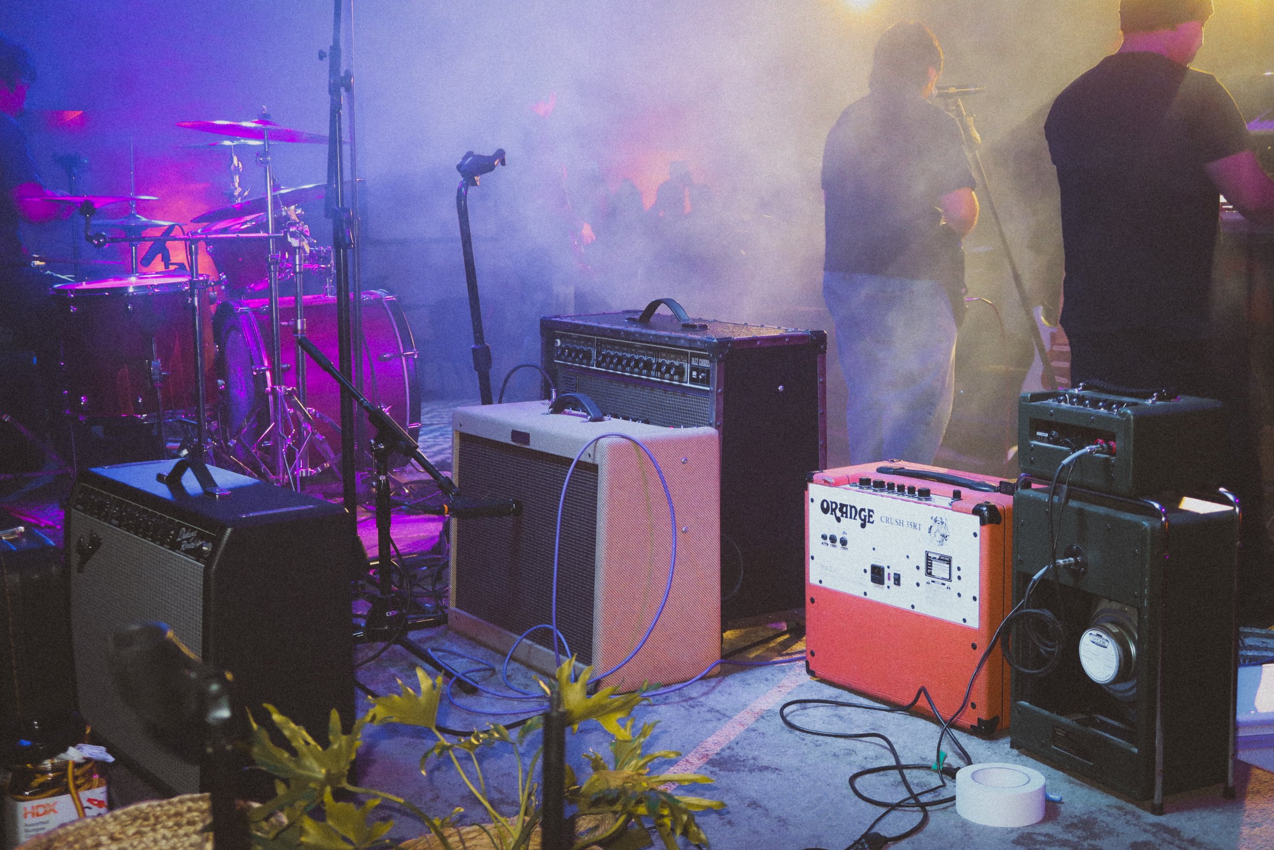 Music stage setup with drums, amplifiers, and musicians in the background, surrounded by colorful stage lighting and fog.